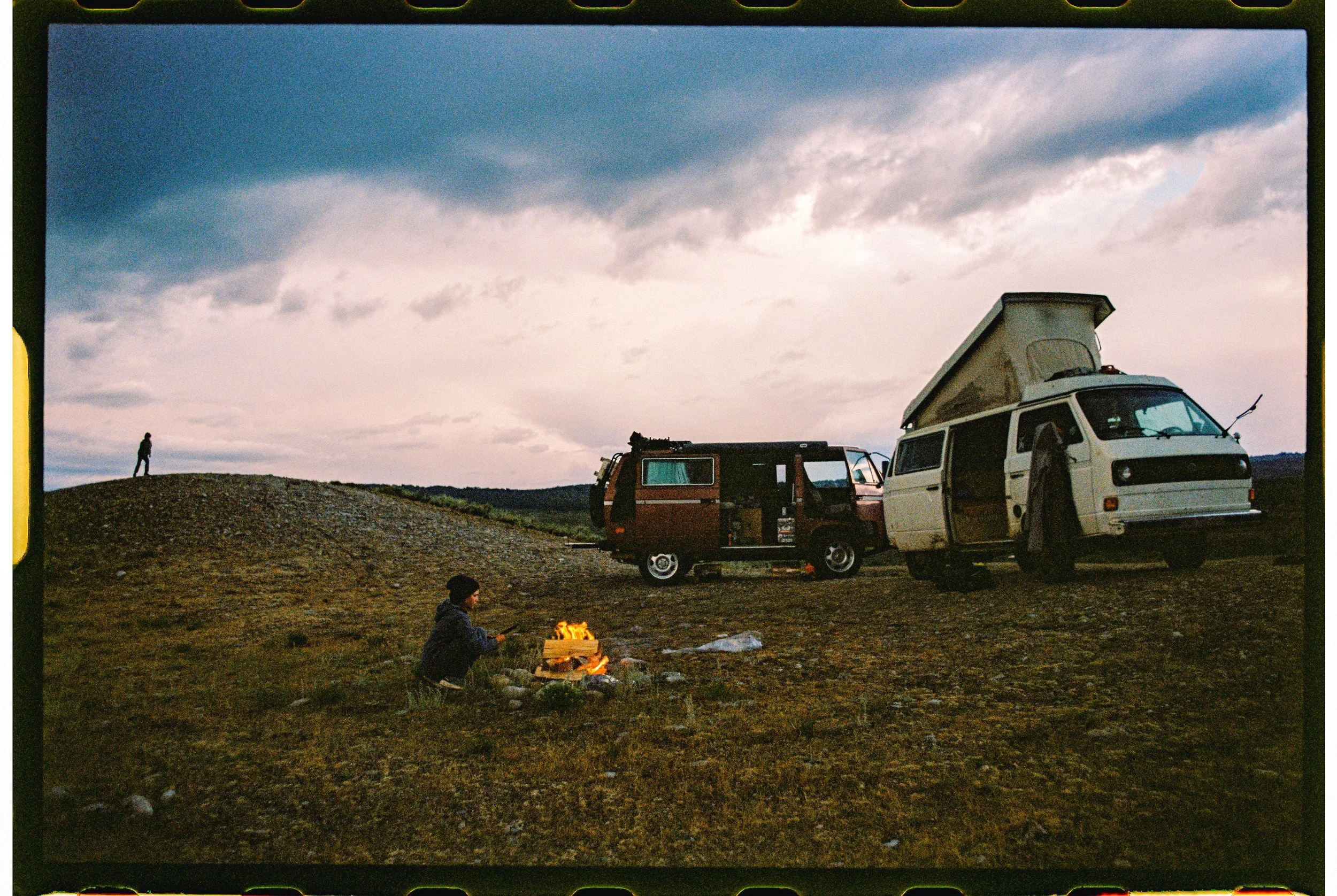 A person sitting by a campfire on a grassy field with two camper vans parked nearby and a person standing on a hill in the background under a cloudy sky.