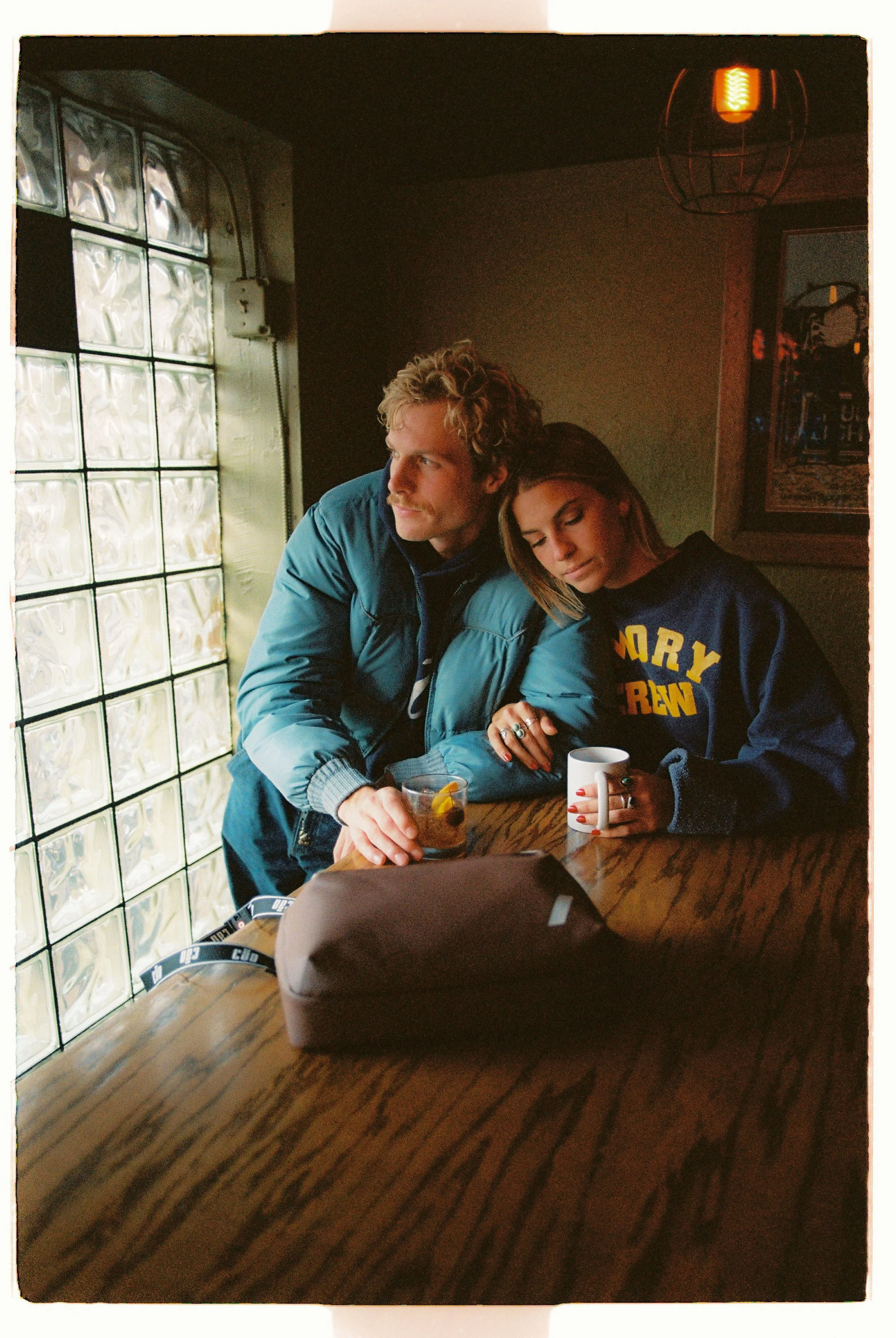 A man and woman sit together at a wooden table in a dimly lit cafe, with the woman resting her head on the man's shoulder. The man holds a glass with a drink, and the woman has a white mug. There are two rings on her fingers, and a brown bag on the table.