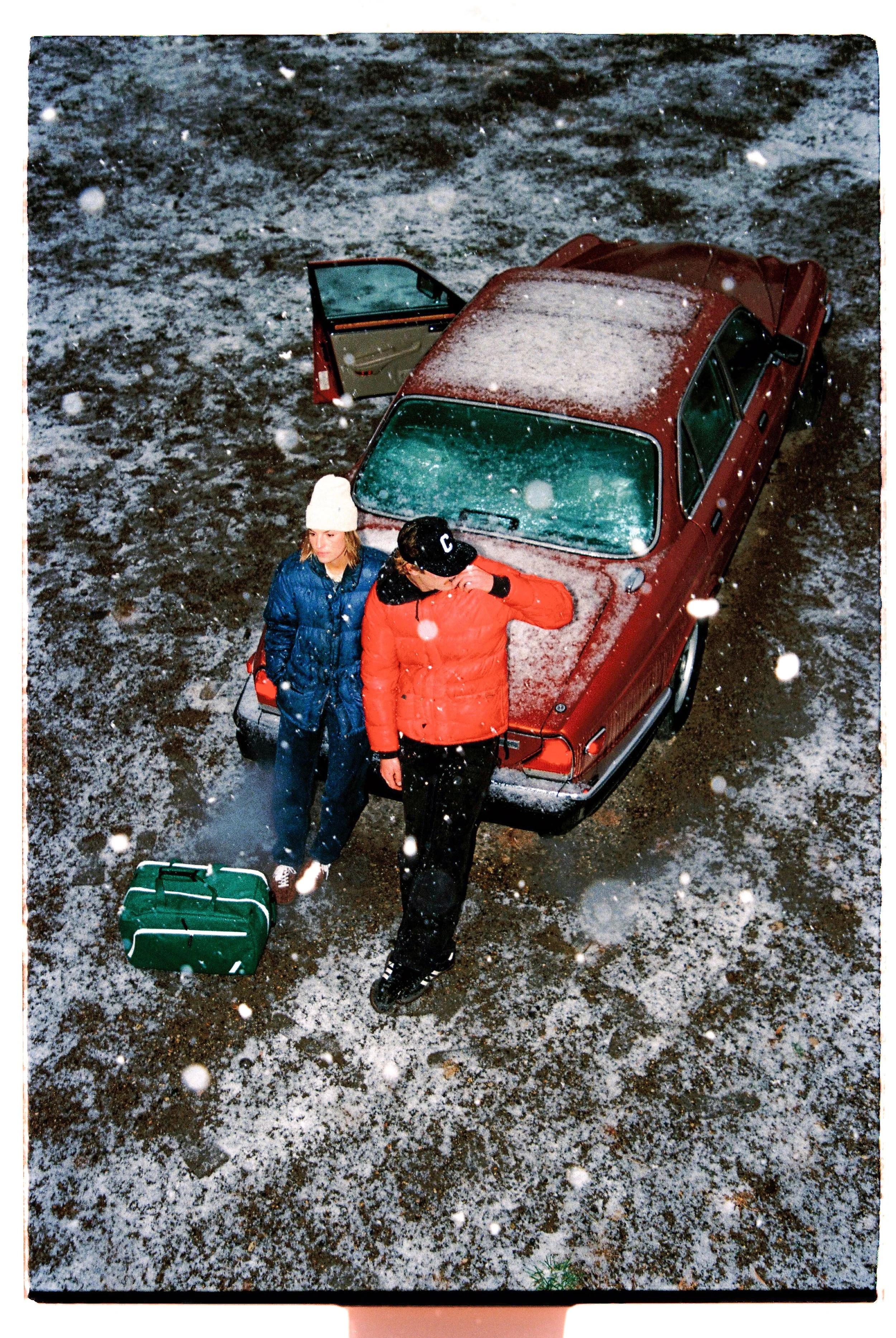 Two people standing next to a red car with a snow-covered roof, on a snowy and icy ground, with a green bag on the ground nearby, during light snowfall.