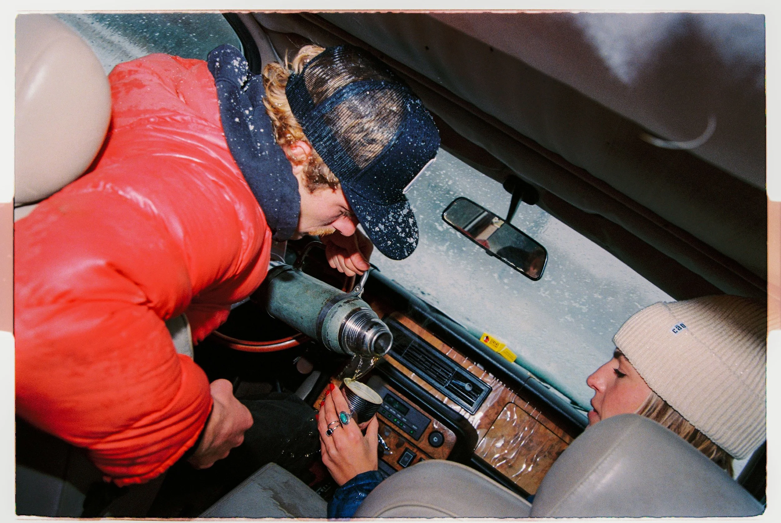 Two people inside a vehicle, with one person pouring a beverage into a cup held by the other. It is raining or snowing outside the vehicle.