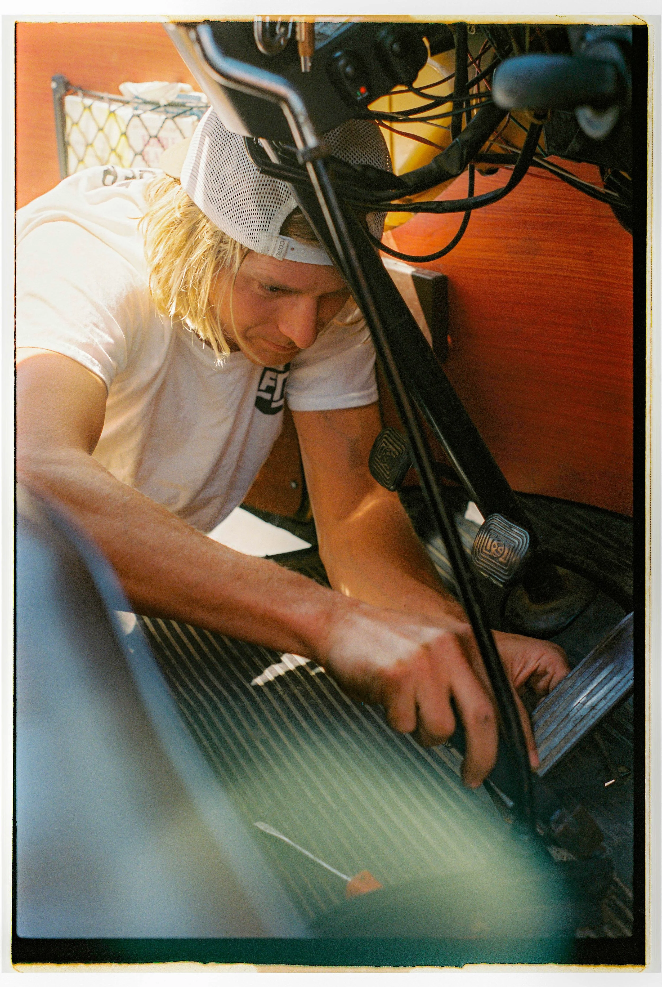 A person with long blonde hair, wearing a white shirt and a gray cap, is working under the dashboard of a vehicle, reaching for the pedals.