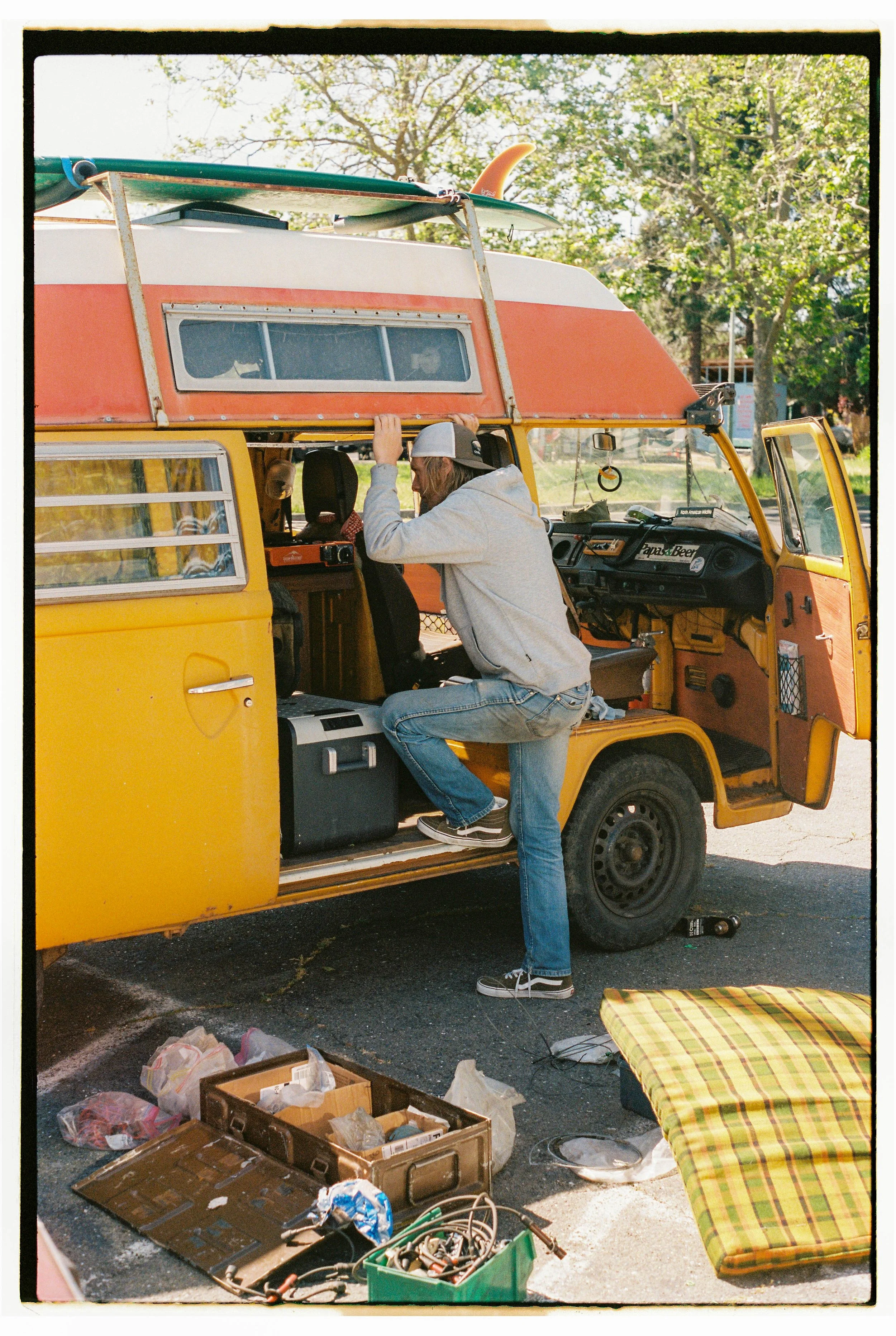 A man working on a yellow and orange camper van parked outdoors, with tools and supplies on the ground around him, and a fishing surfboard on the roof.