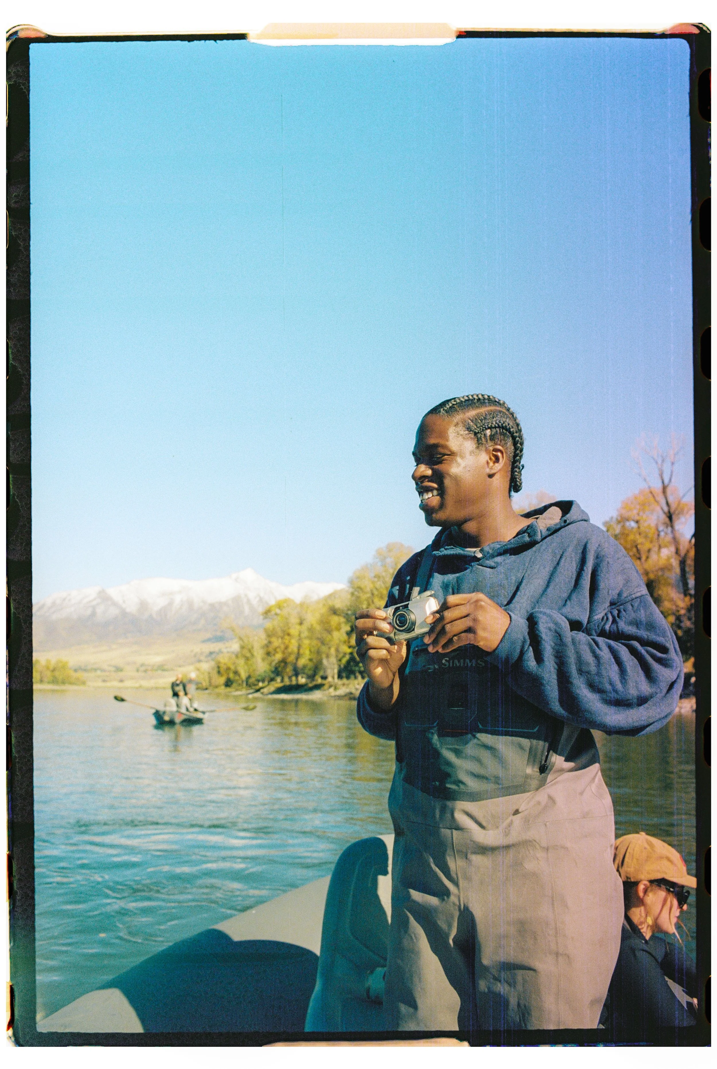 A person standing on a boat holding a camera, with mountains, water, and trees in the background, during a clear day.