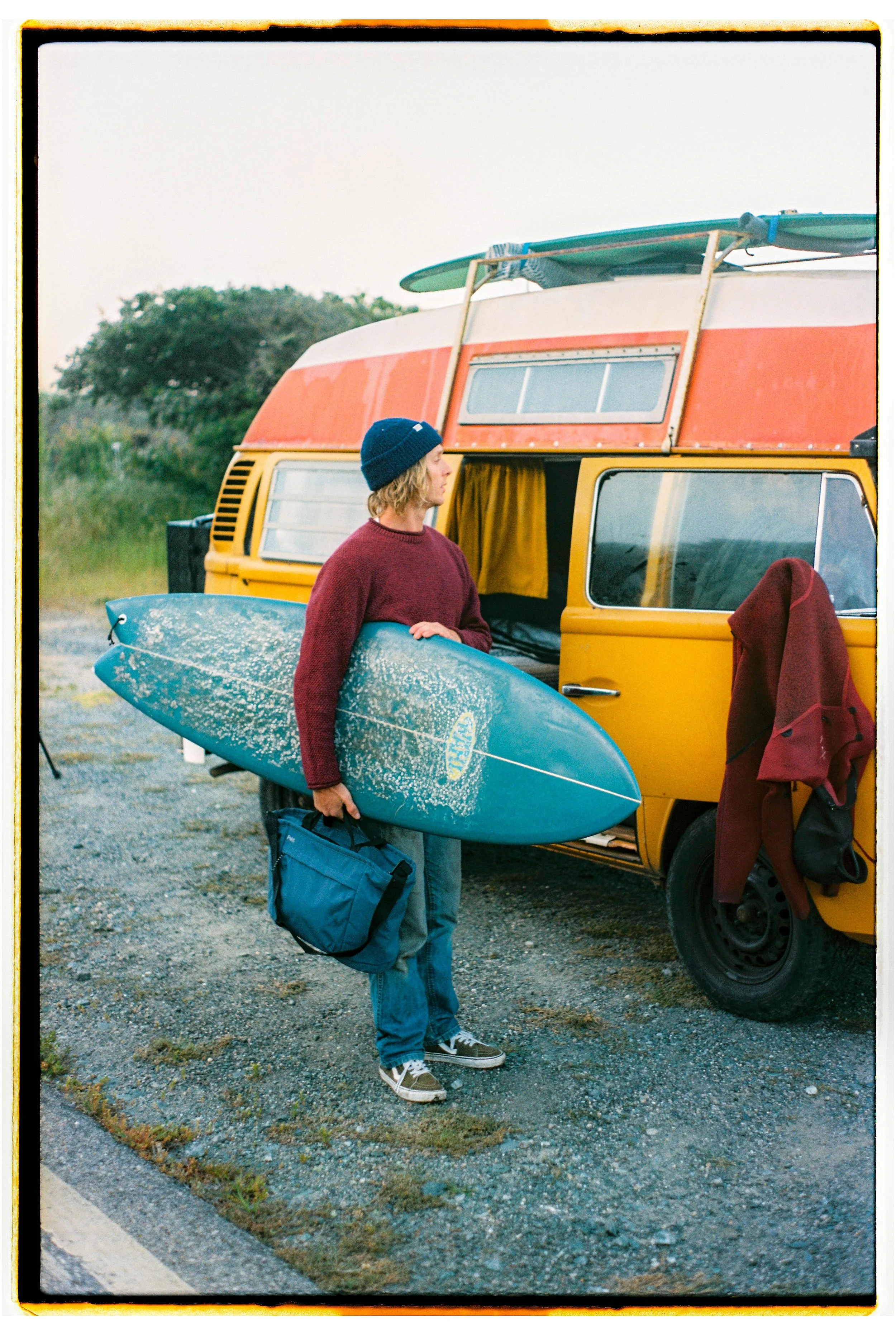 A person with long hair, wearing a blue beanie, maroon sweater, jeans, and sneakers, holding a blue surfboard and a blue bag, standing next to a yellow and red camper van with a surfboard on top. A red jacket hangs on the door handle.