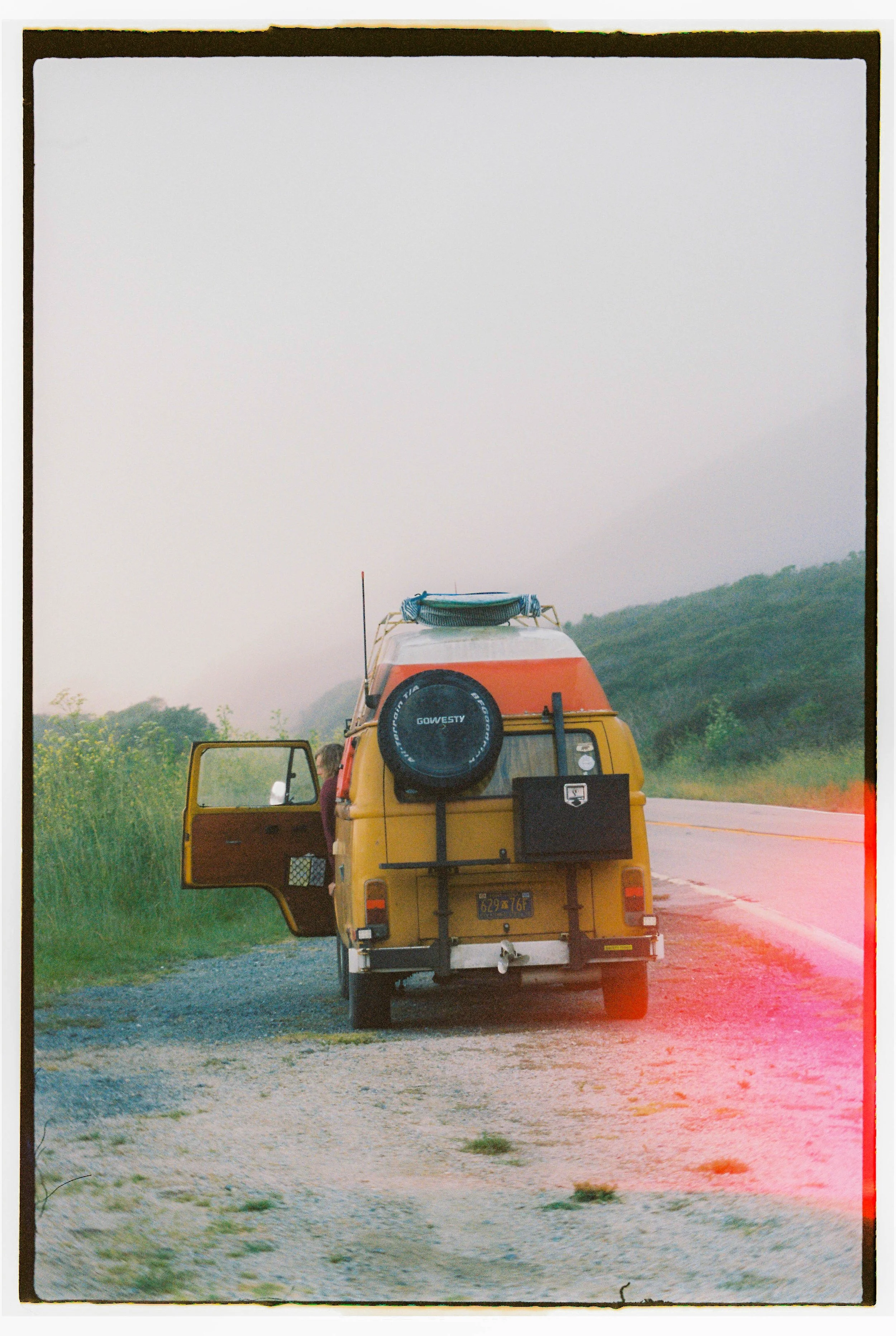 Rear view of a yellow camper van parked on a dirt area beside a road, with a person standing by the open door, surrounded by green hills and a foggy sky.