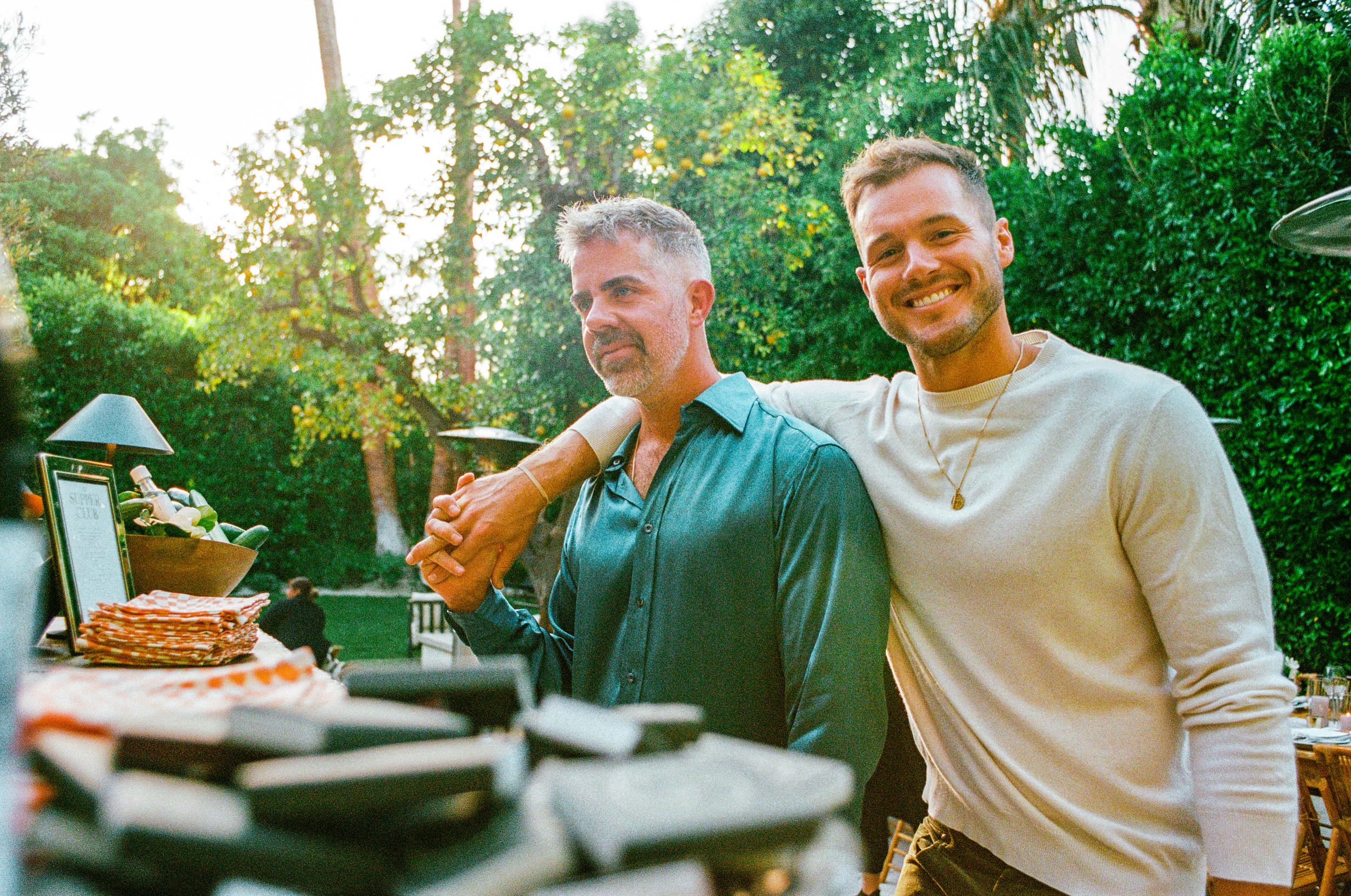 Two men at an outdoor gathering, one with gray hair in a teal shirt, and the other with brown hair in a cream sweater, smiling and standing close together.
