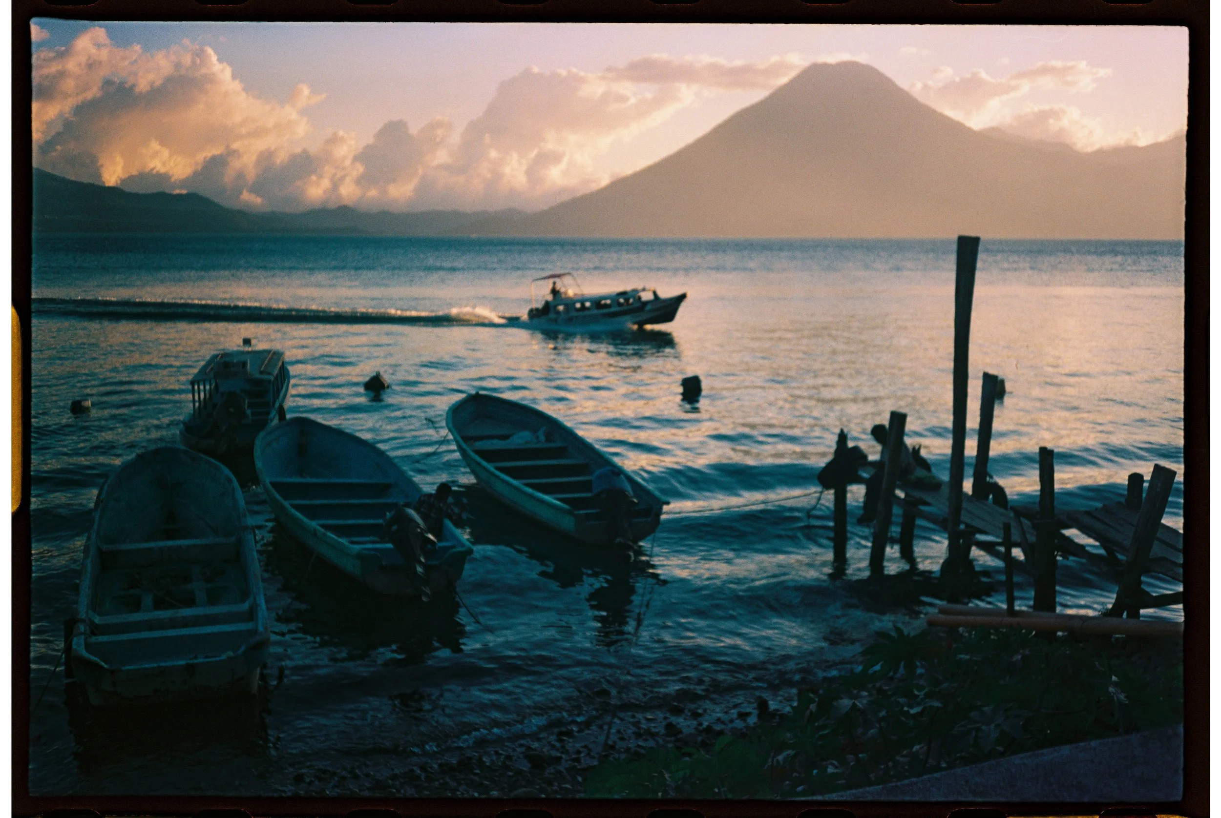 Boats docked near a pier on a lake with a volcano in the background, under a partly cloudy sky during sunset.