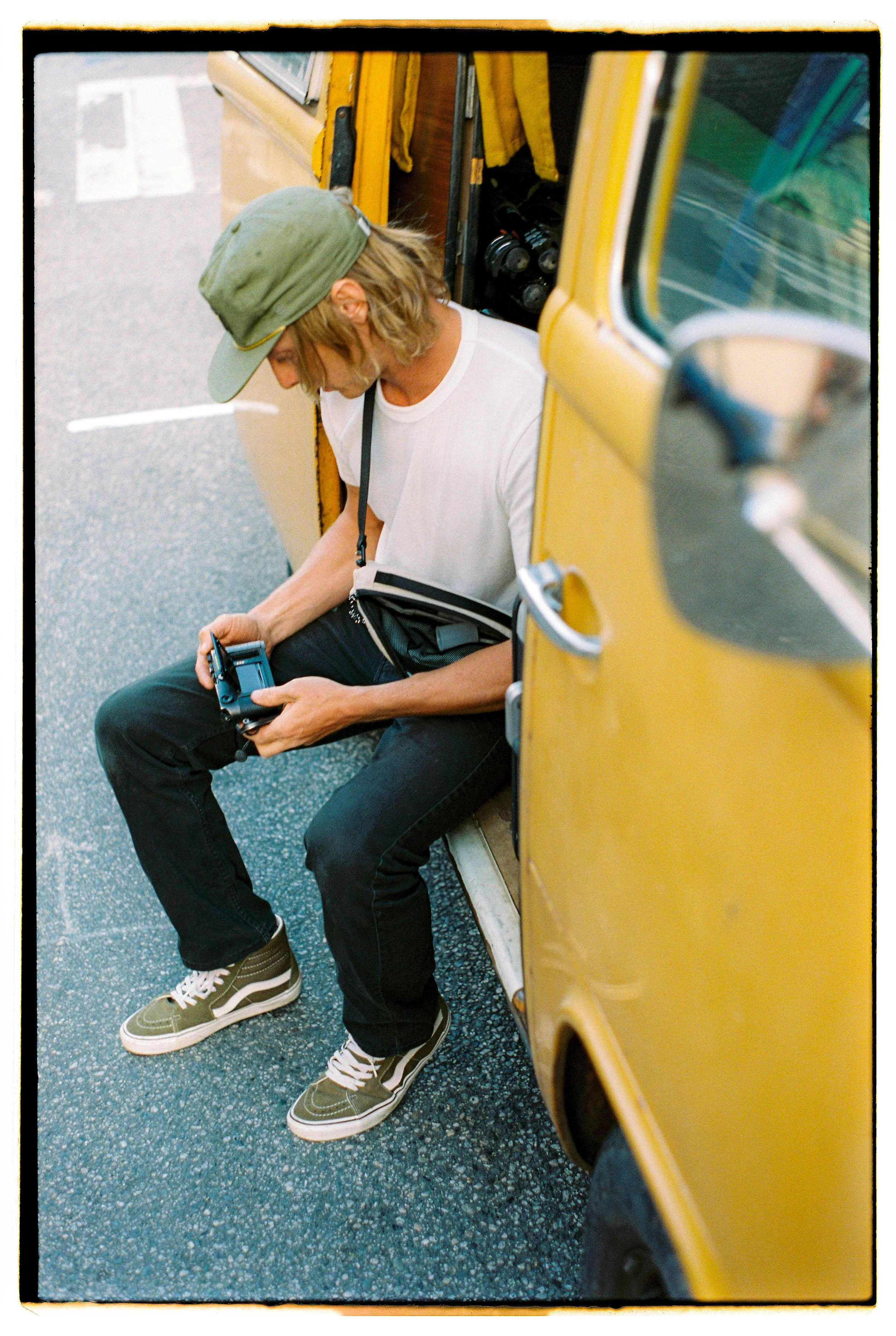 Person sitting on the street next to a yellow three-wheeled vehicle, looking at a device in their hands, wearing a green cap, white t-shirt, black pants, and sneakers.