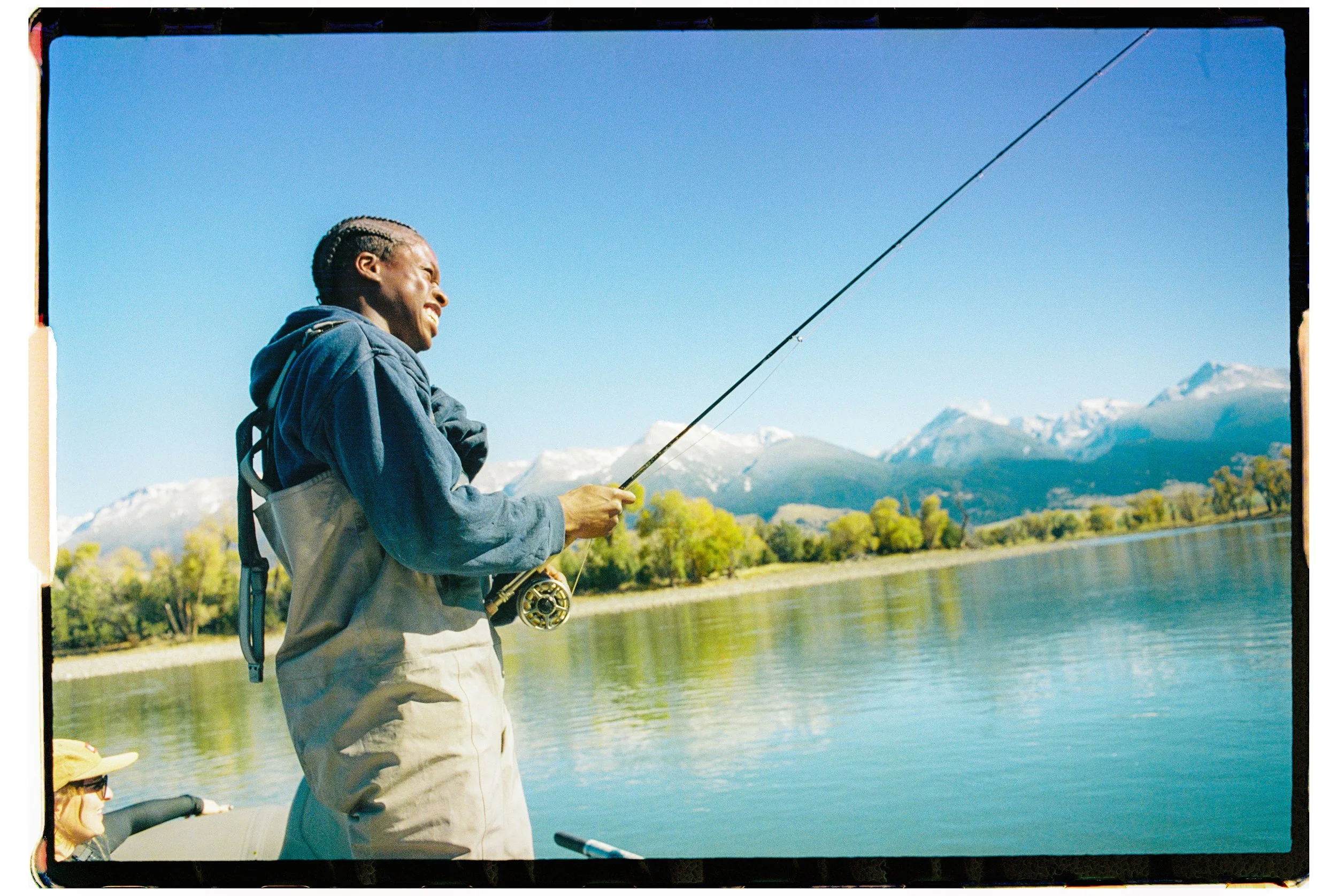 Person smiling and fishing by a lake with mountains and trees in the background on a sunny day.