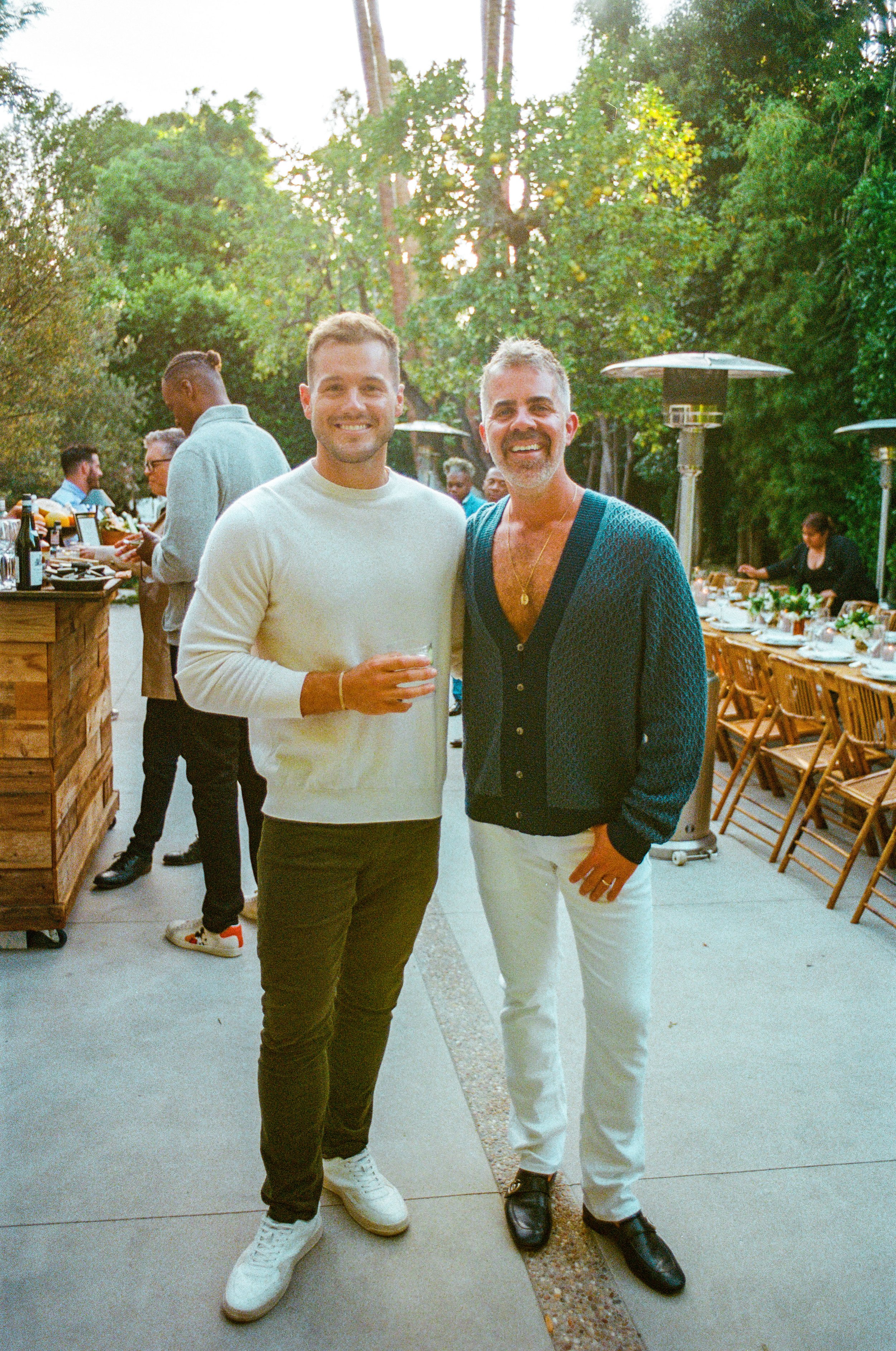 Two men smiling and posing for a photo at an outdoor gathering during daytime, with tables set for a meal and other guests in the background.