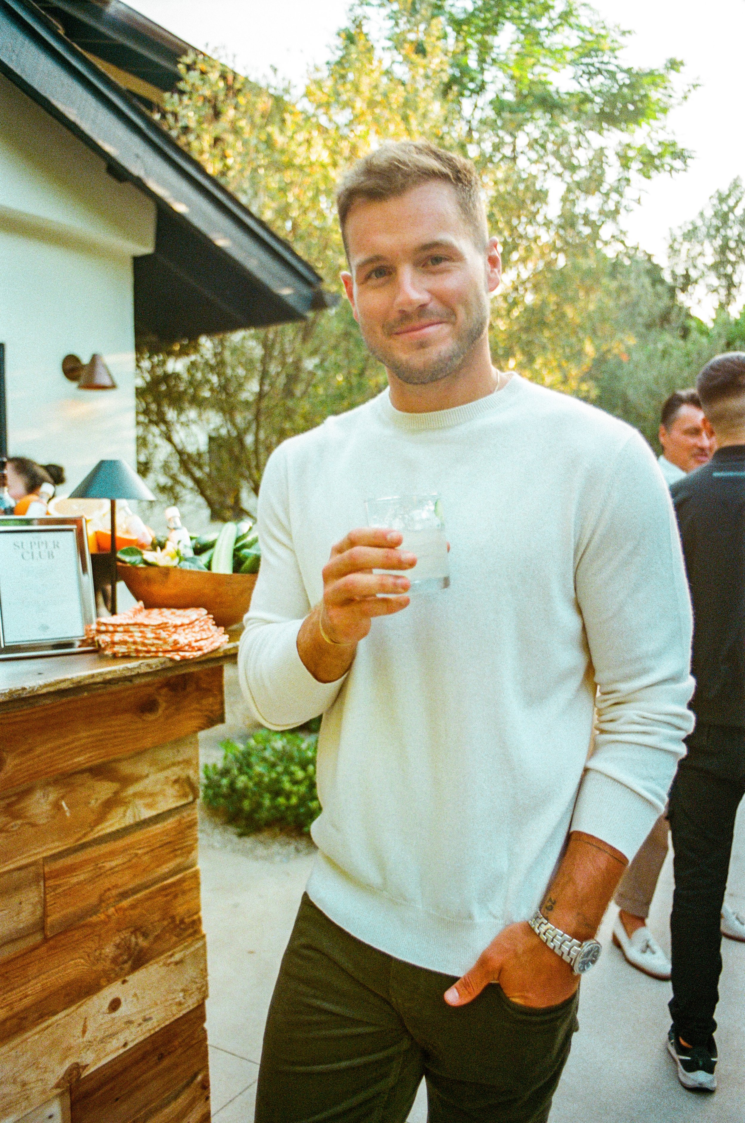 A man in a white sweater holding a drink at an outdoor event, with a rustic bar and other people in the background.