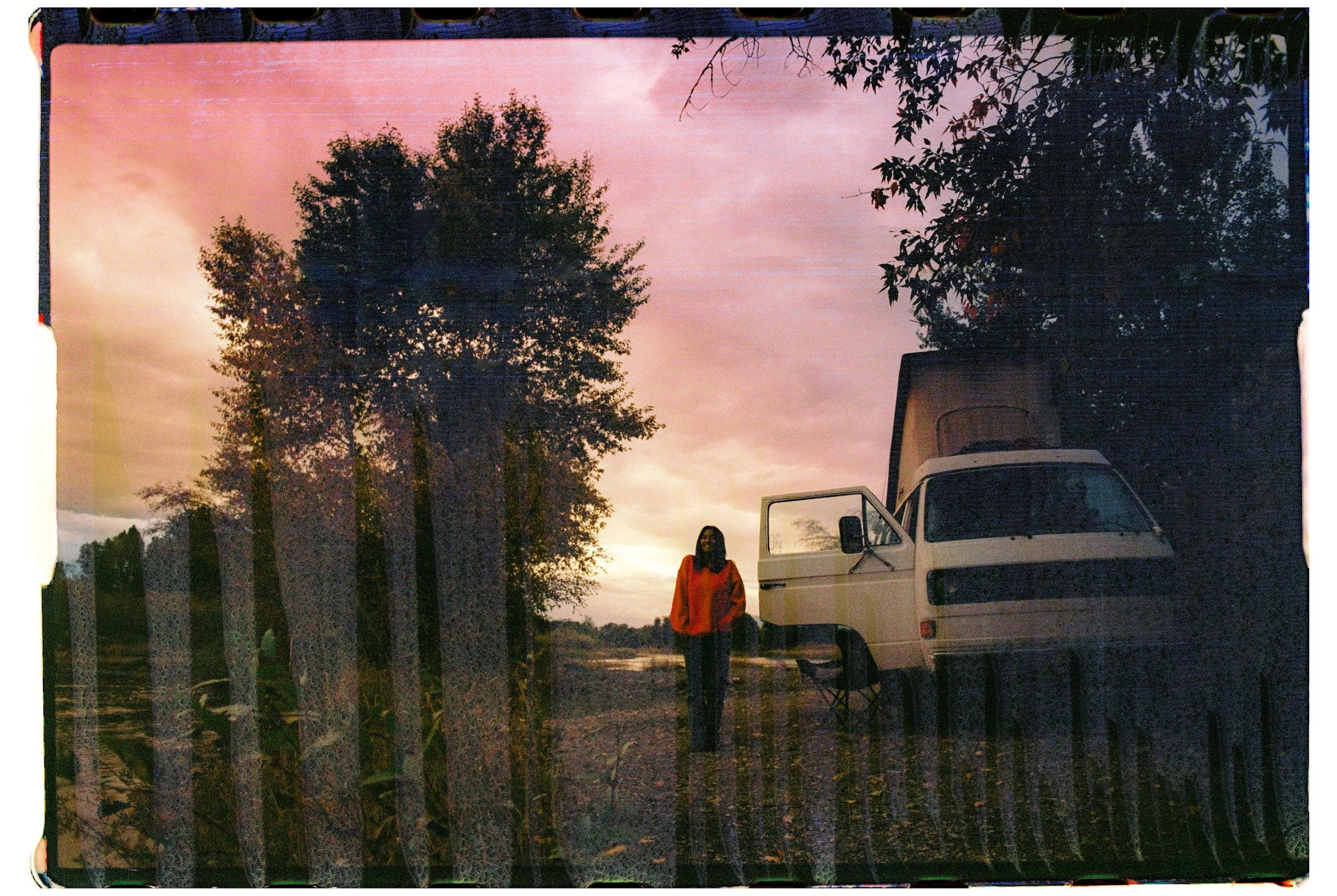 A person standing outside near a white van with an open door during sunset, with tall trees and a fence in the foreground, and pink and orange sky in the background.