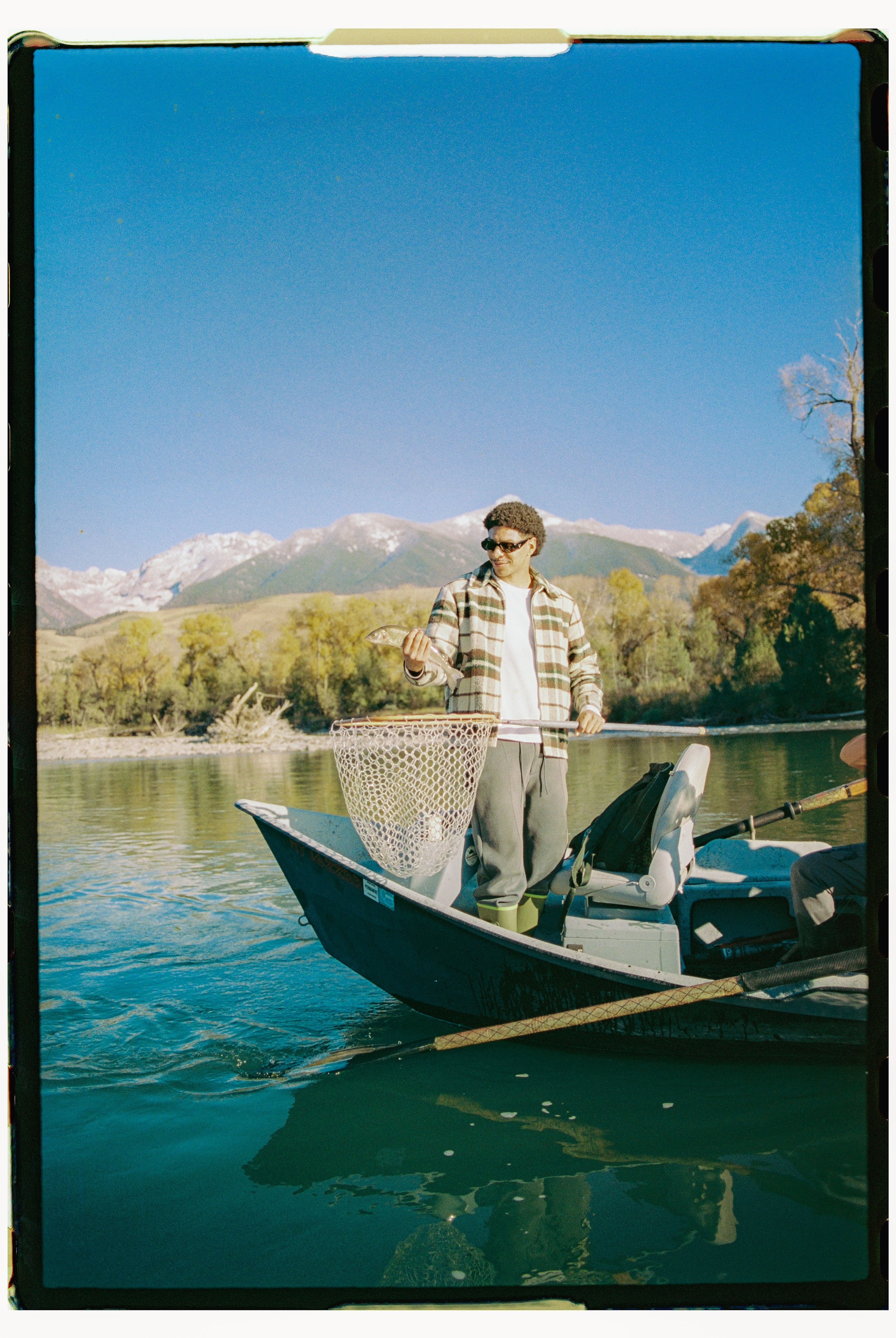 A man standing on a small boat fishing in a calm lake, with mountains and trees in the background.