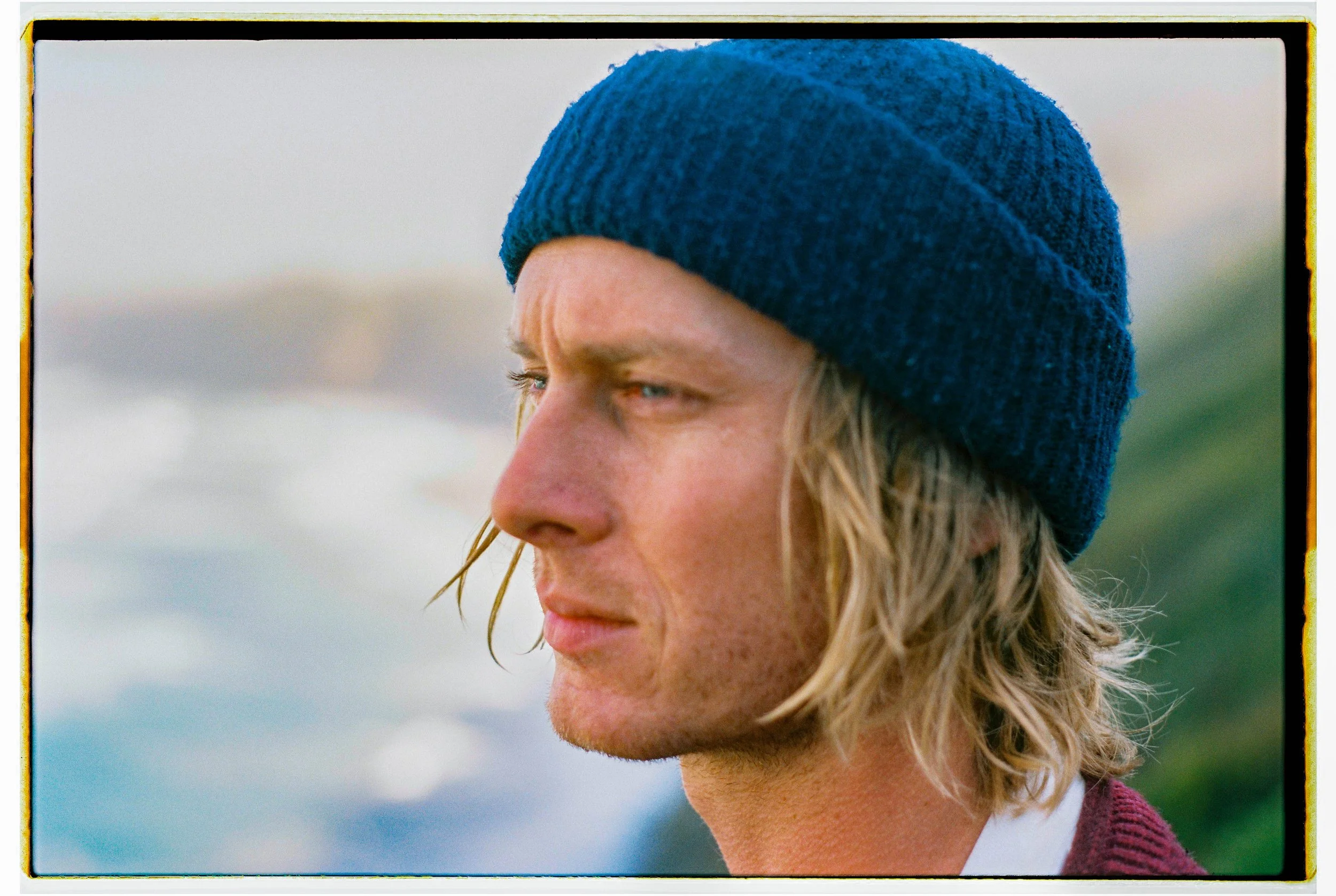 Close-up of a young man with long blonde hair wearing a blue beanie, looking thoughtfully into the distance outdoors.
