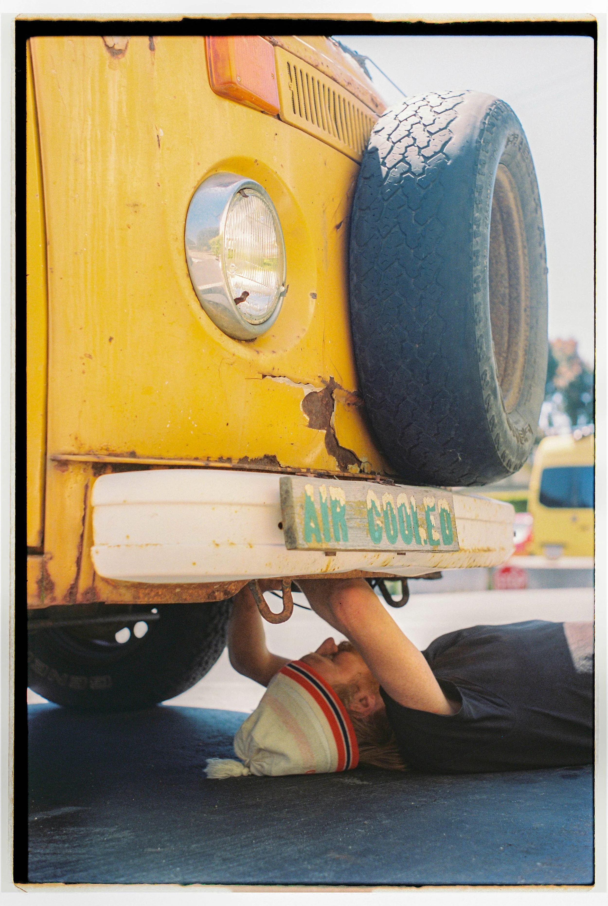A person lying on the ground under the front of a yellow vintage truck, working on it. The truck has a spare tire mounted on the front and a cracked paint job. The license plate reads 'AIR COOLED'.