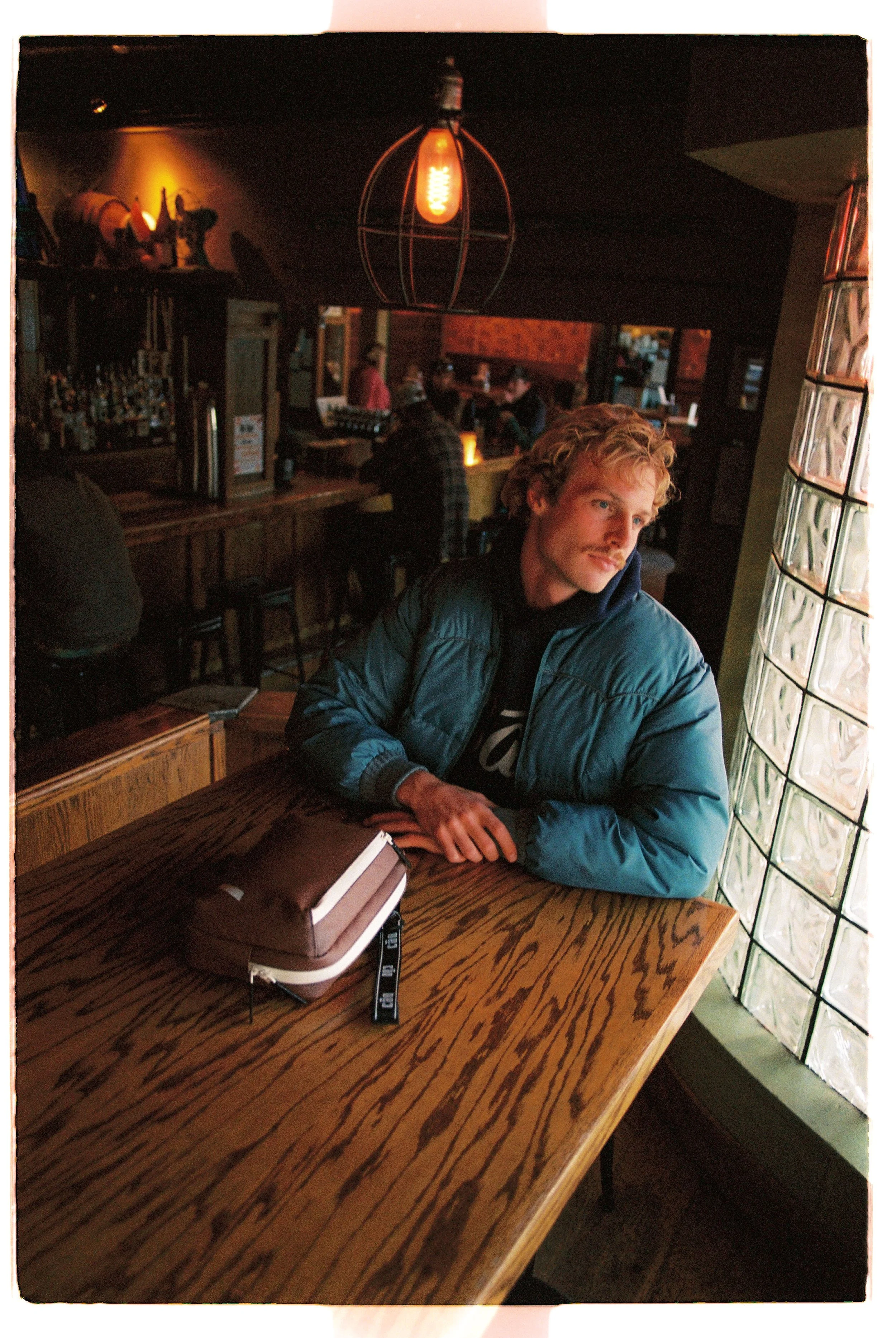 A young man with blond, curly hair and light skin sitting alone at a wooden table in a dimly lit bar or restaurant, wearing a blue puffer jacket and looking out of the window to his right.