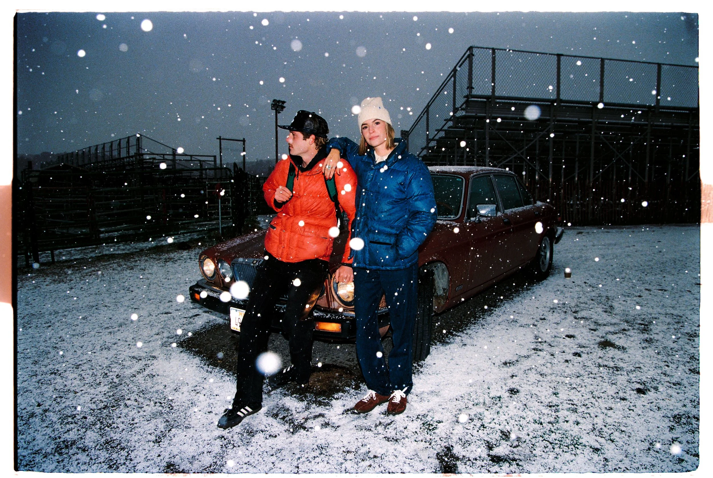 Two young people standing outdoors in a snowy field near a vintage car, wearing winter clothing, with snow falling and a cloudy sky overhead.