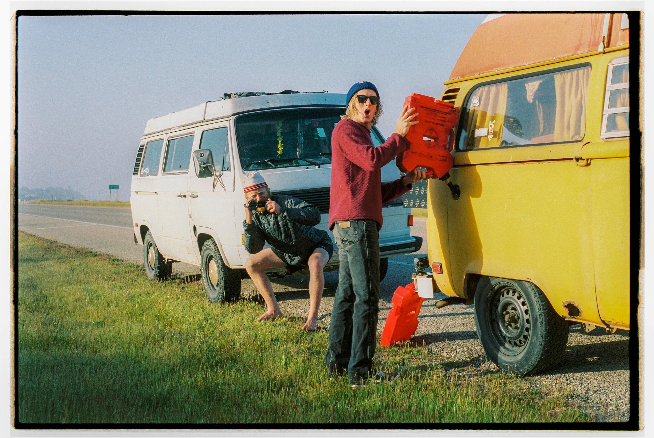 Two young men on a roadside, one crouching with a camera and the other holding a red fuel can poured into a yellow vintage camper trailer.