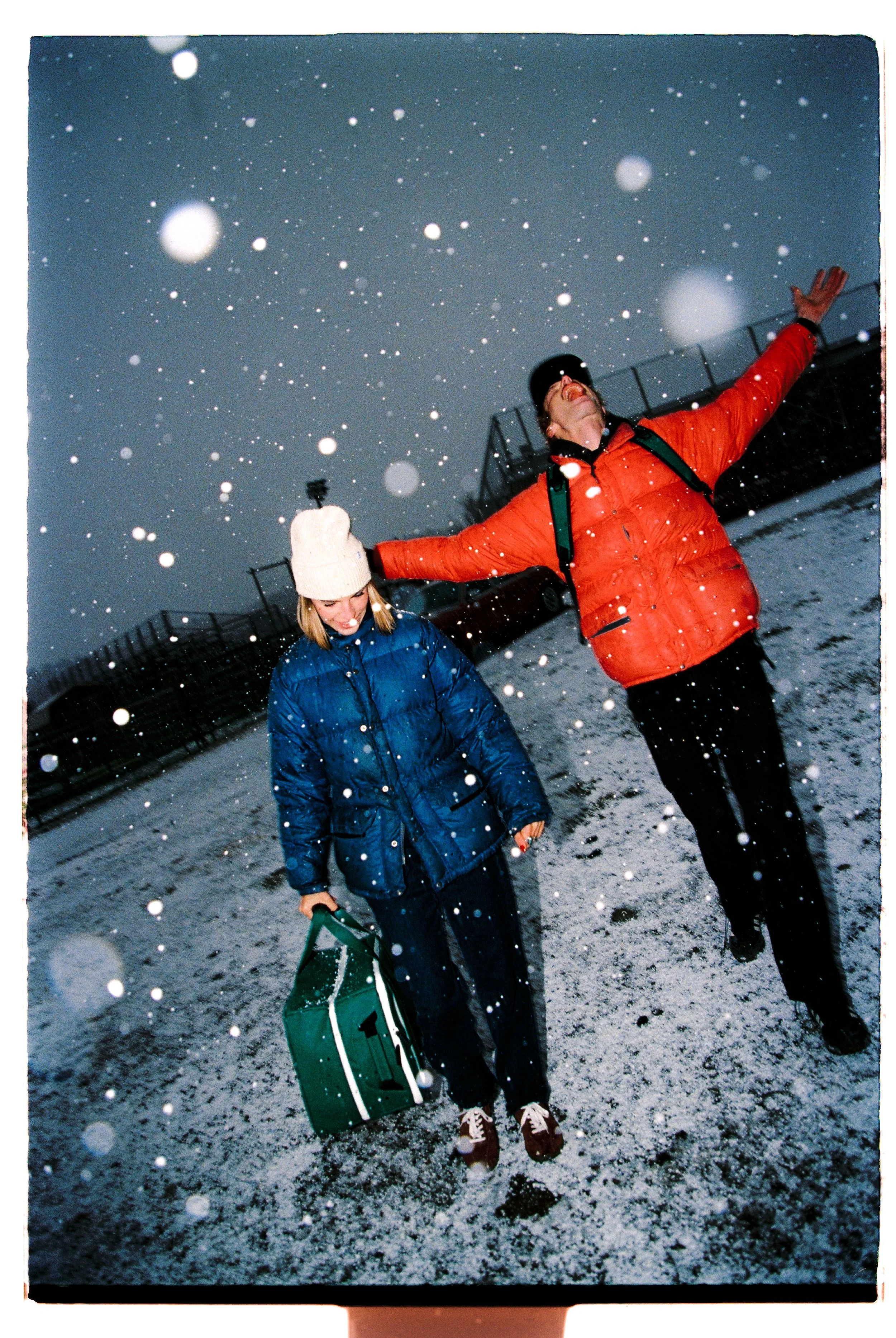 A woman and a man are standing outside in the snow, smiling, with their arms outstretched. The woman is holding a green bag and wearing a white knit hat and a blue puffy jacket. The man is wearing a red puffy jacket and a black cap. Snow is falling h