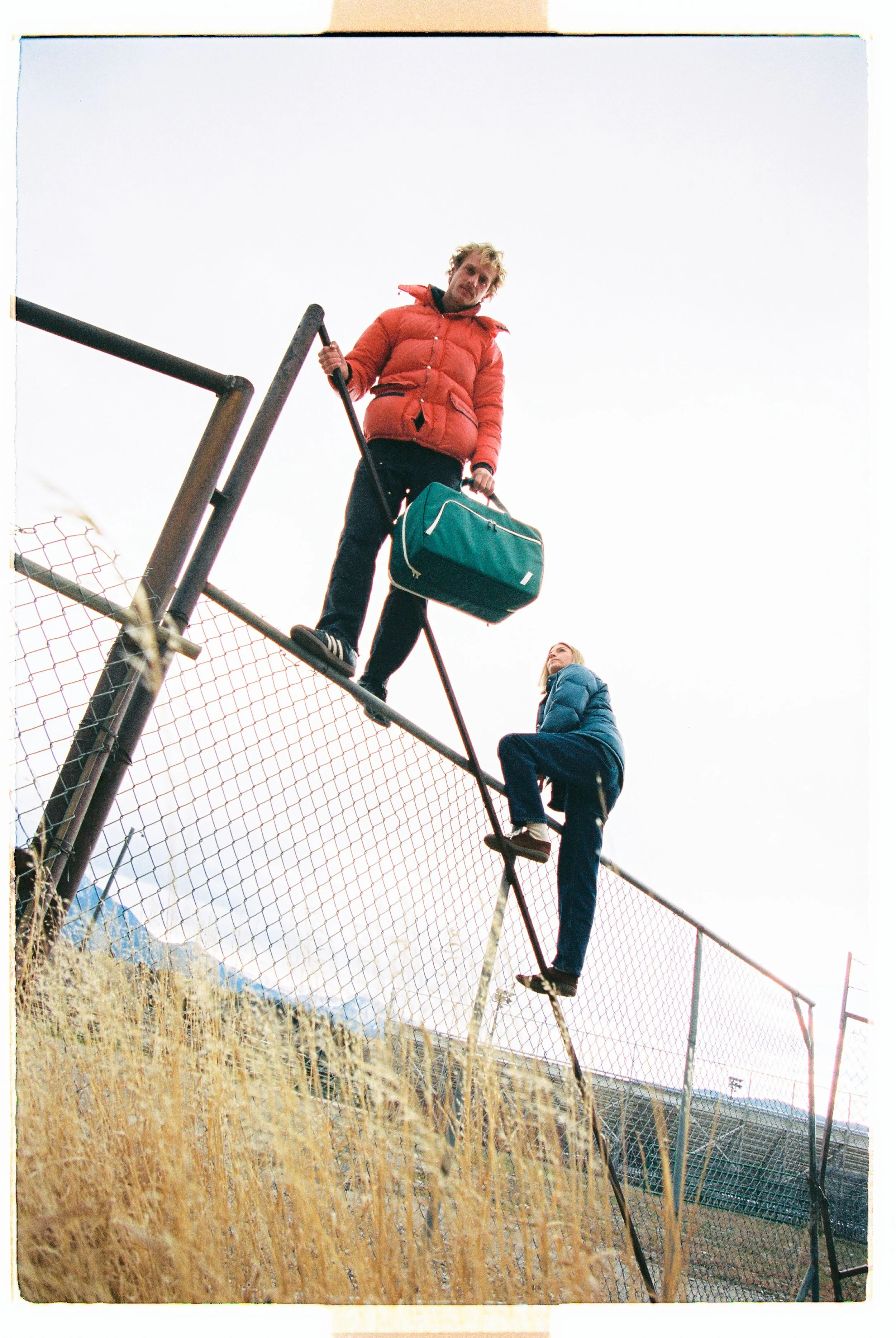 Two people on top of a chain-link fence, one holding a duffel bag and the other climbing behind, in a grassy area under an overcast sky.