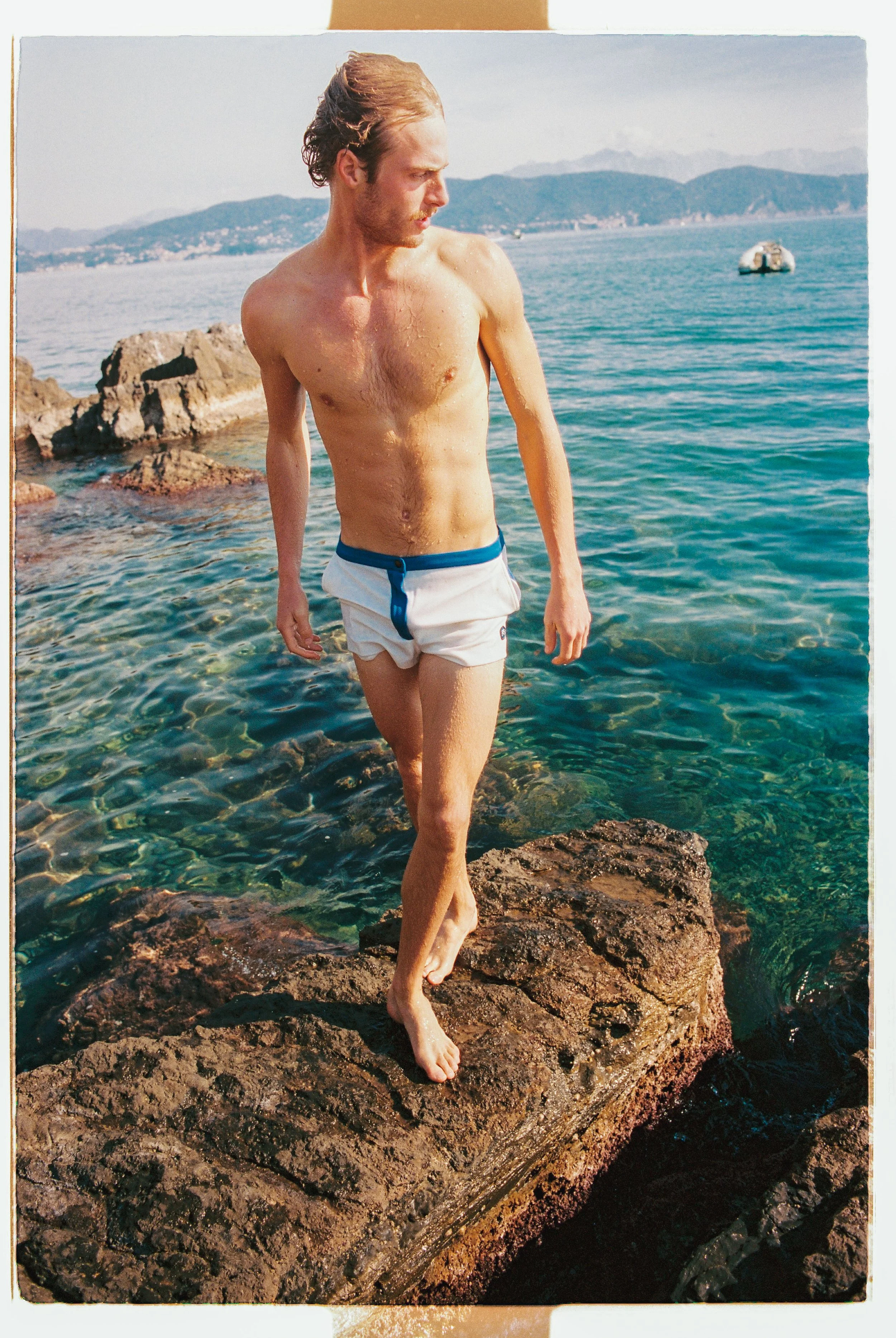 A man with long hair and a beard is walking barefoot on rocks near water, wearing white swim trunks with a blue waistband, with mountains in the background.