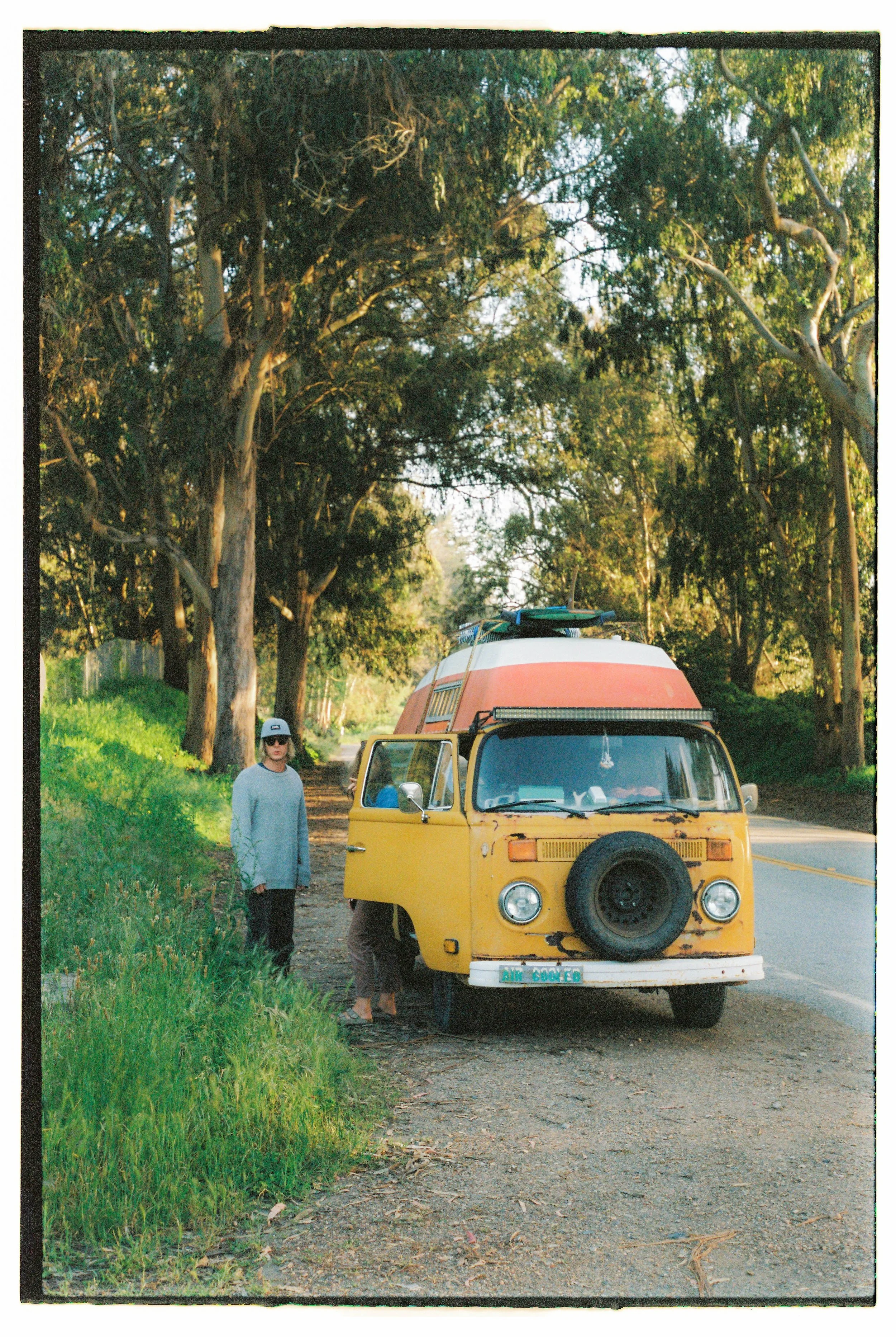 A person standing next to a vintage yellow van with a surfboard on top, parked beside a tree-lined road in a rural area.