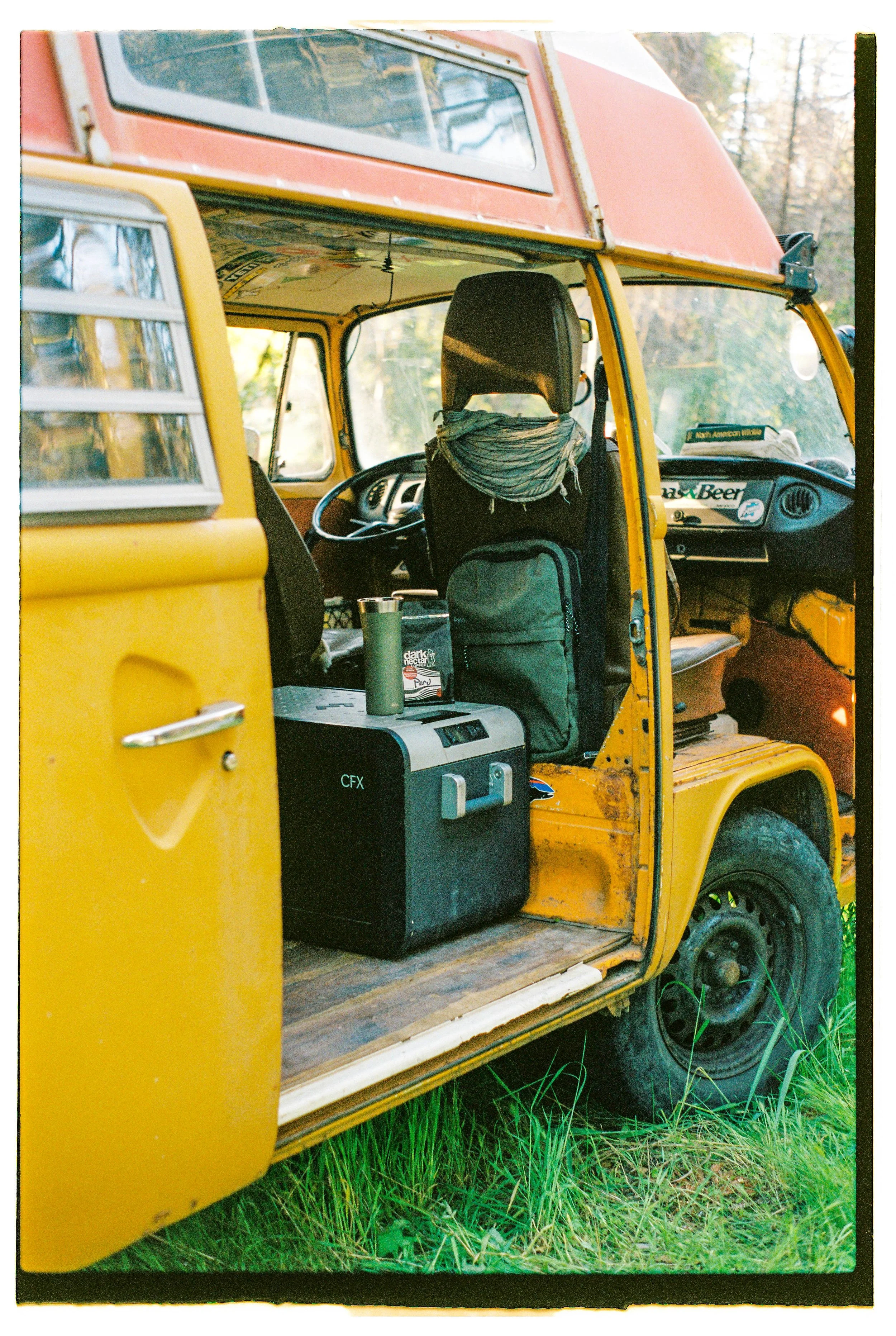 Interior of a yellow vintage van with items such as a backpack, a cooler, and a water bottle inside, parked on grass with trees in the background.