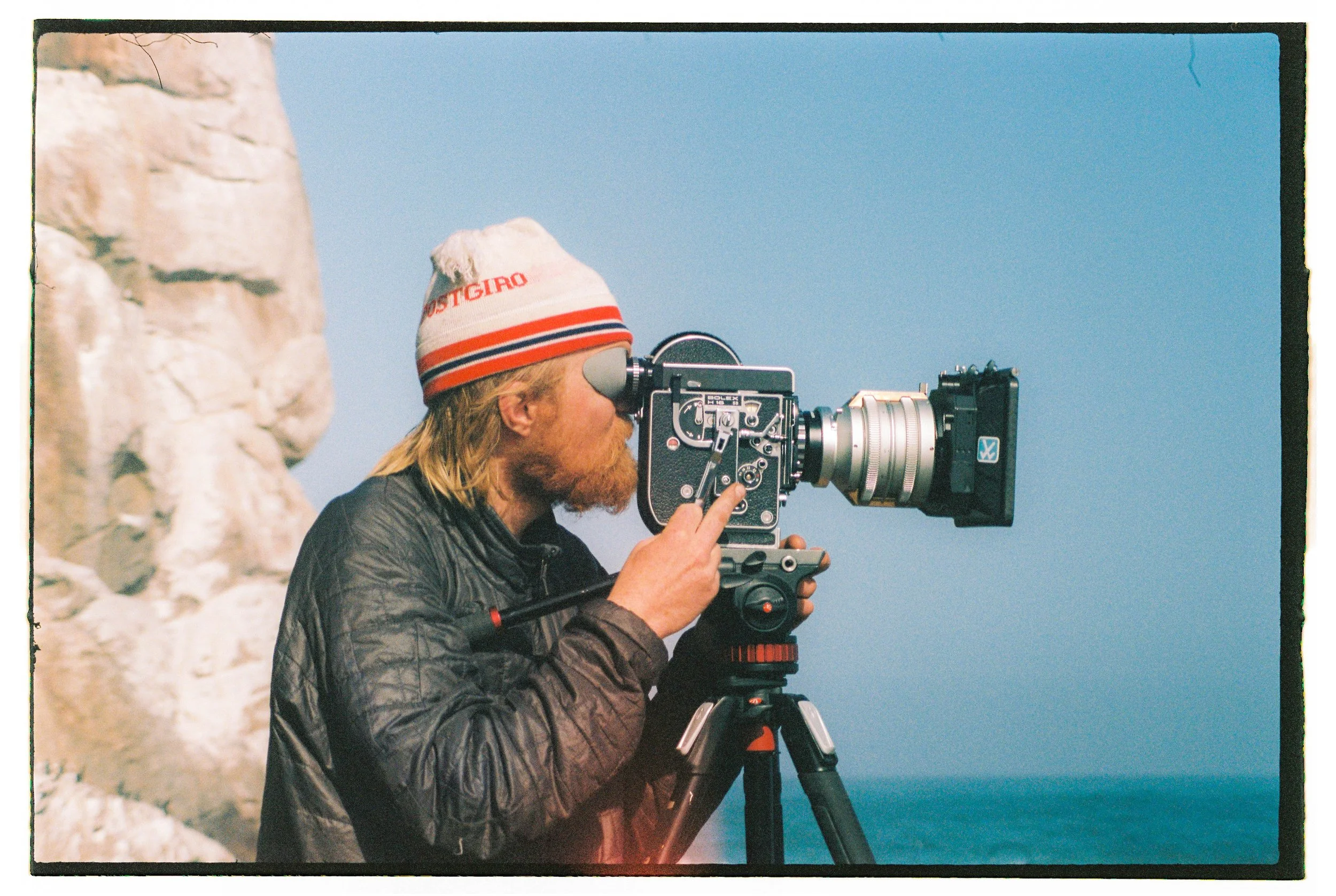 A person with long blond hair, a red beard, wearing a white beanie with red and blue stripes, looks through a film camera mounted on a tripod, with a backdrop of blue sky and a rocky cliff by the ocean.