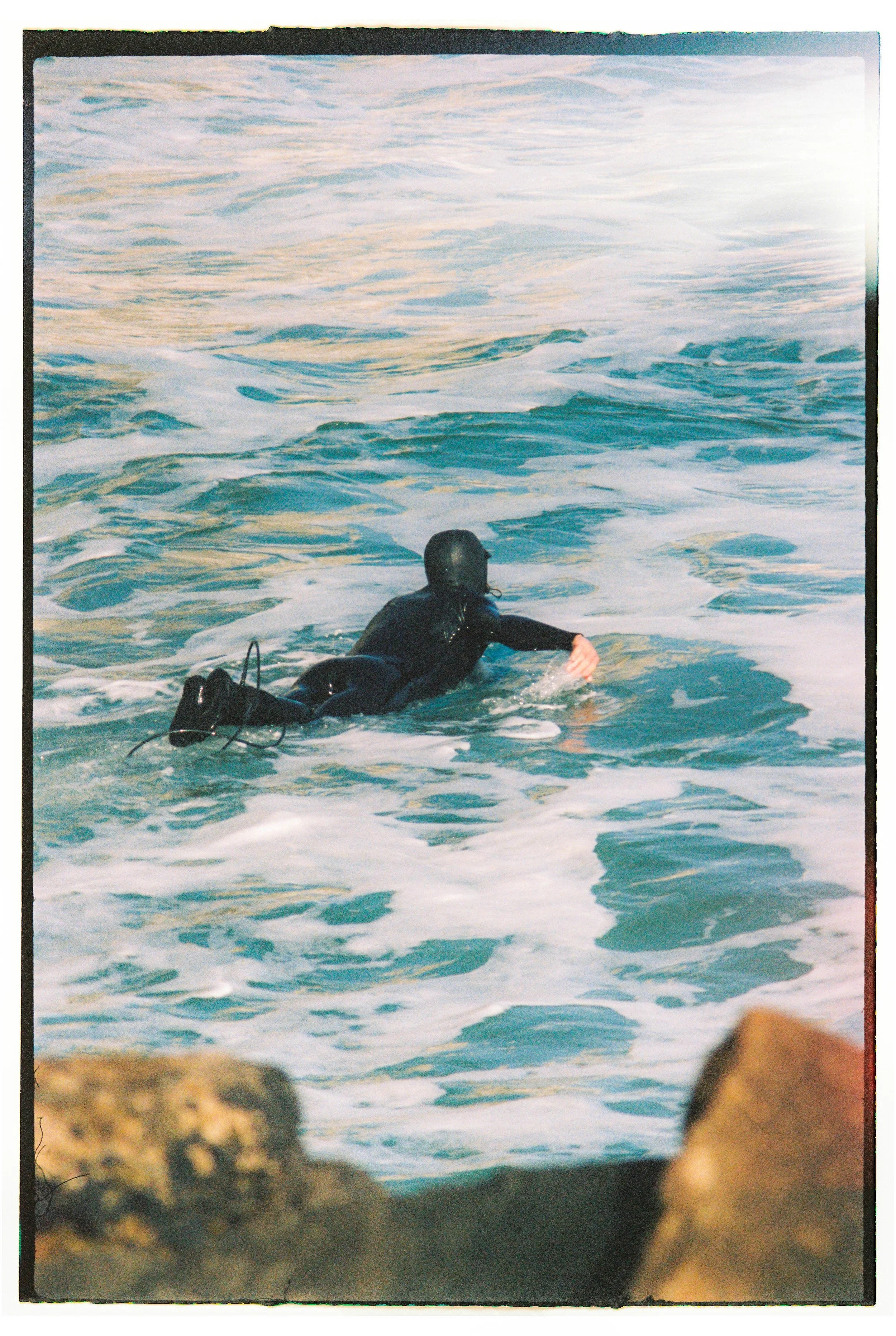 A person in a black wetsuit and diving mask swimming in the ocean near rocks.