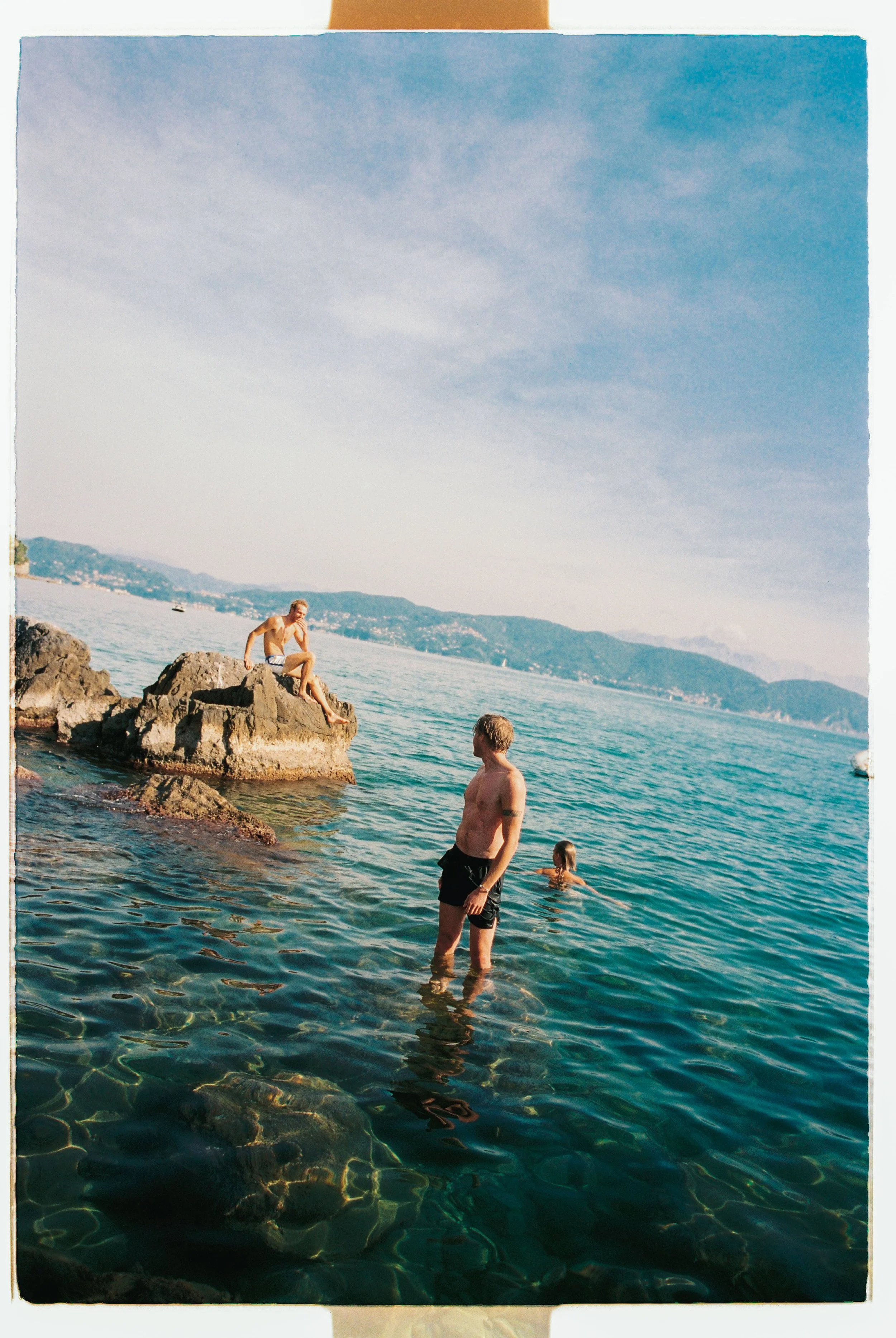 People swimming and relaxing in clear water near rocks on a beach with mountains in the background.