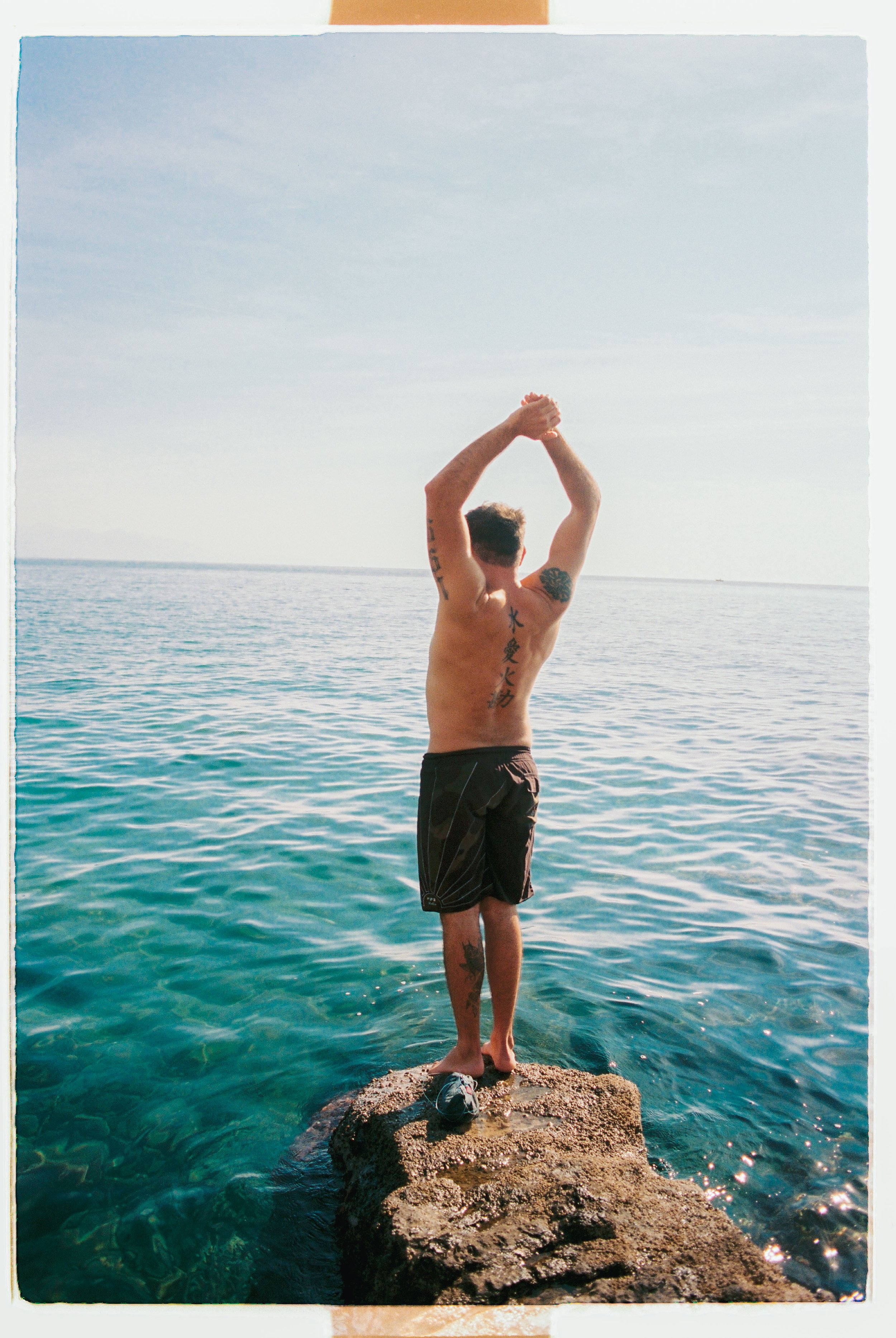 Young man standing on a rock by the water, facing away, stretching his arms above his head near the ocean under a light blue sky.