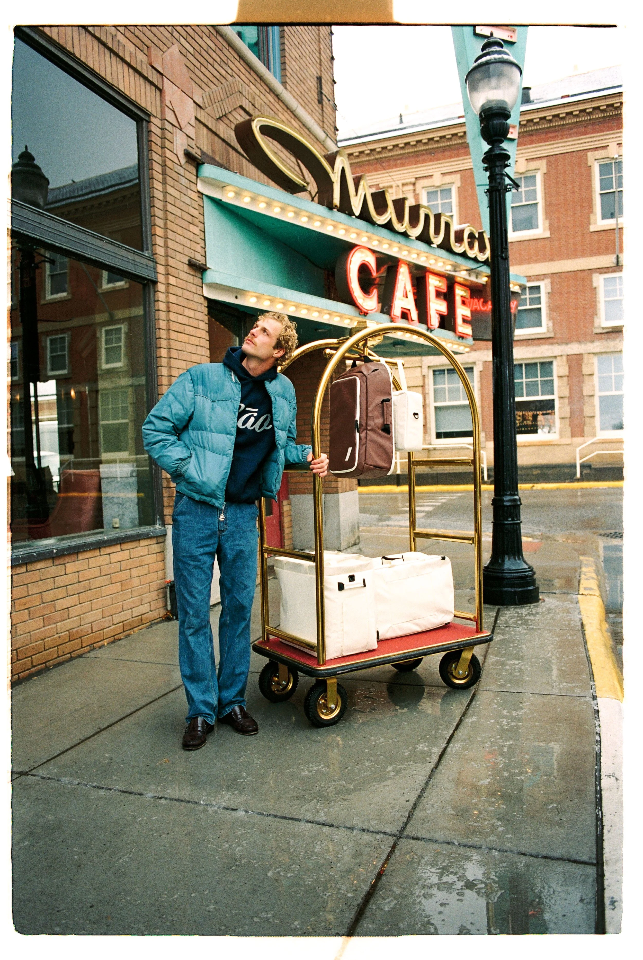 A young man stands outside a cafe with a luggage cart, holding the handle, with backpacks and bags on it. The cafe has a vintage marquee sign and a brick exterior. It is raining, and the man wears a blue jacket and jeans, looking up.