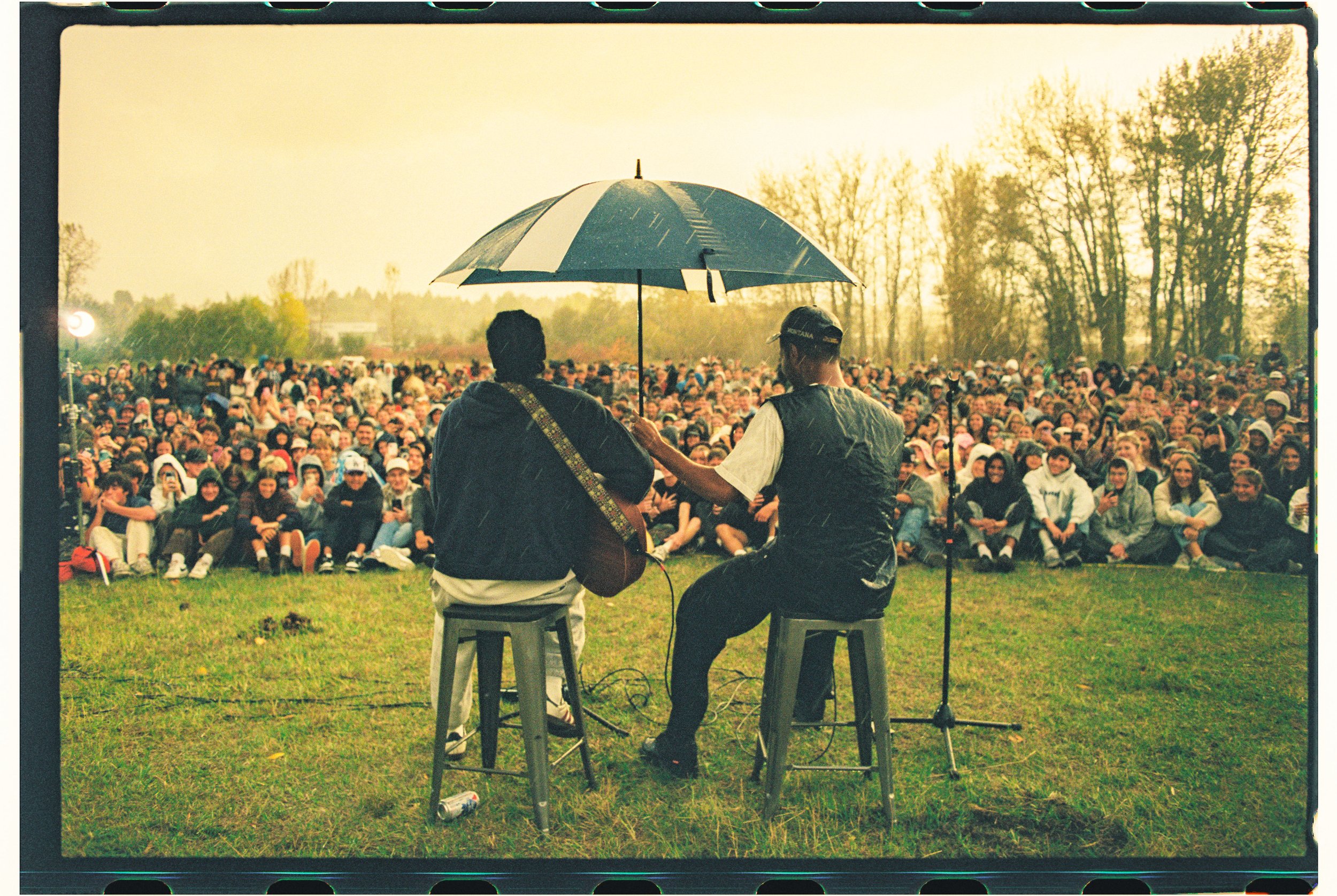 Two musicians performing on stage outdoors in front of a large seated crowd during the daytime. One musician is playing guitar, and both are sitting on backless stools. There is an umbrella over them, and a microphone stand is visible.