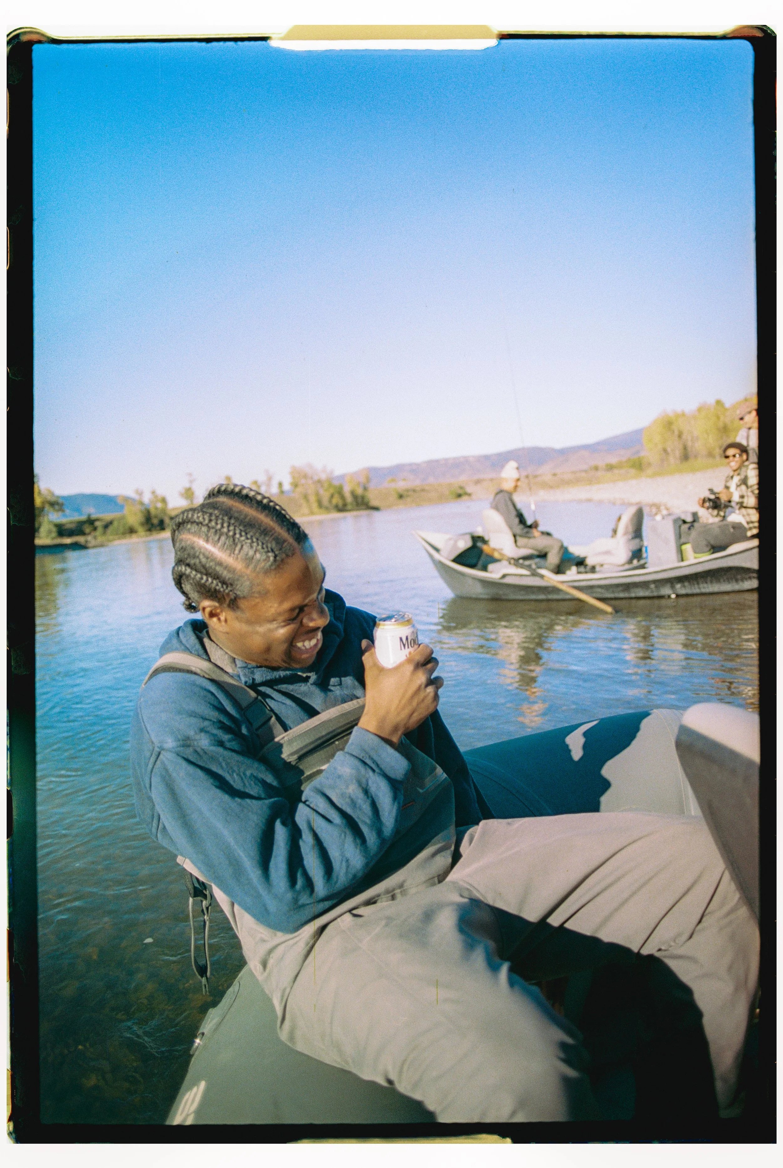 Person sitting on the side of a boat, smiling and laughing while holding a can of soda, with a calm river and another small boat with people fishing in the background.