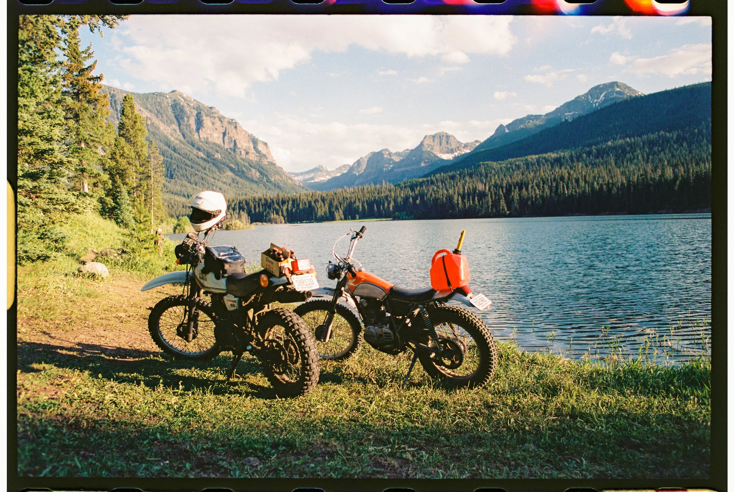 Two vintage motorcycles parked by a lake with mountains and trees in the background, under a partly cloudy sky.