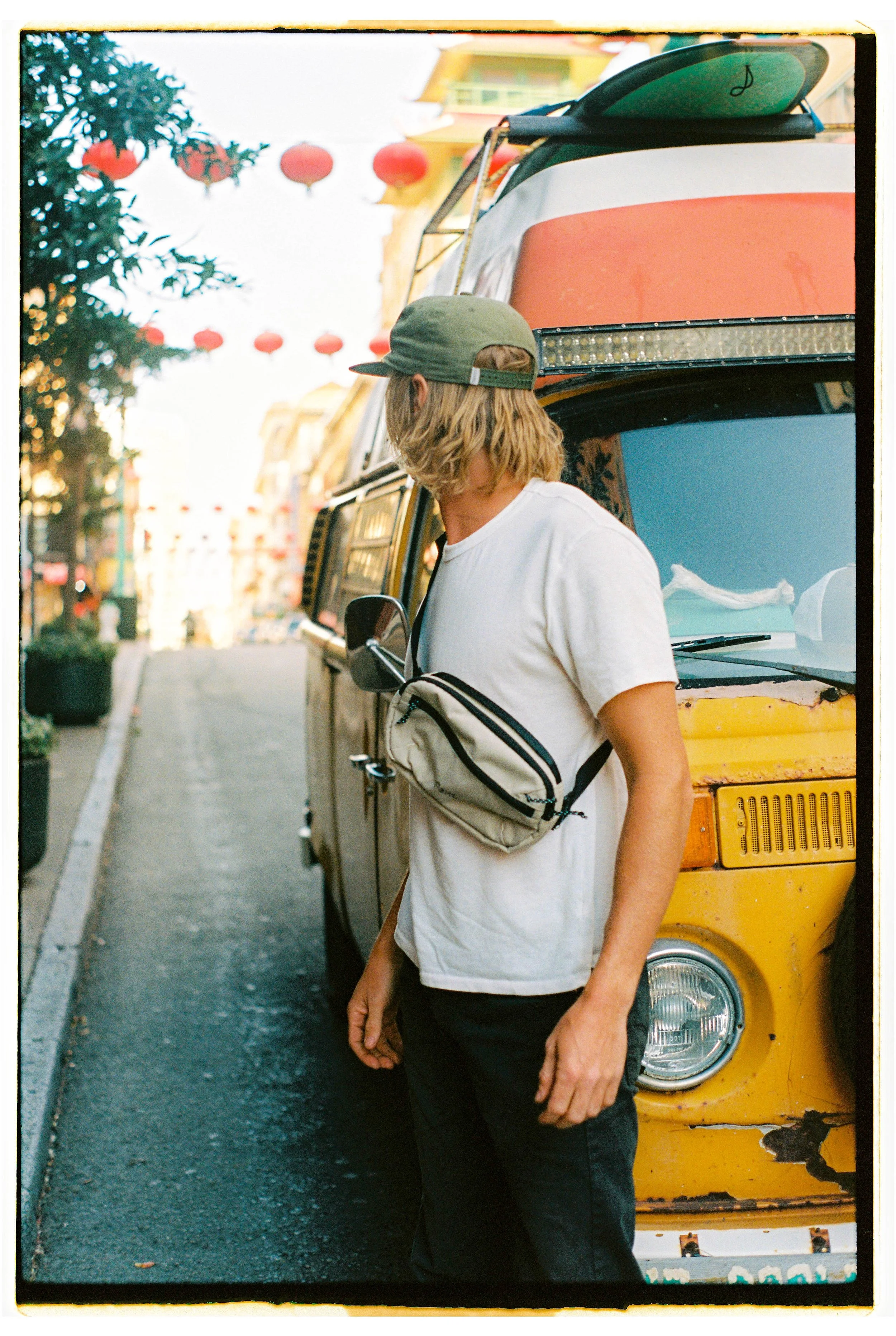 A young man with shoulder-length blonde hair, wearing a green cap, white t-shirt, black pants, and a crossbody bag, stands on a city street next to a yellow vintage van with a red and white surfboard on top. Red lanterns hang above the street, sugges