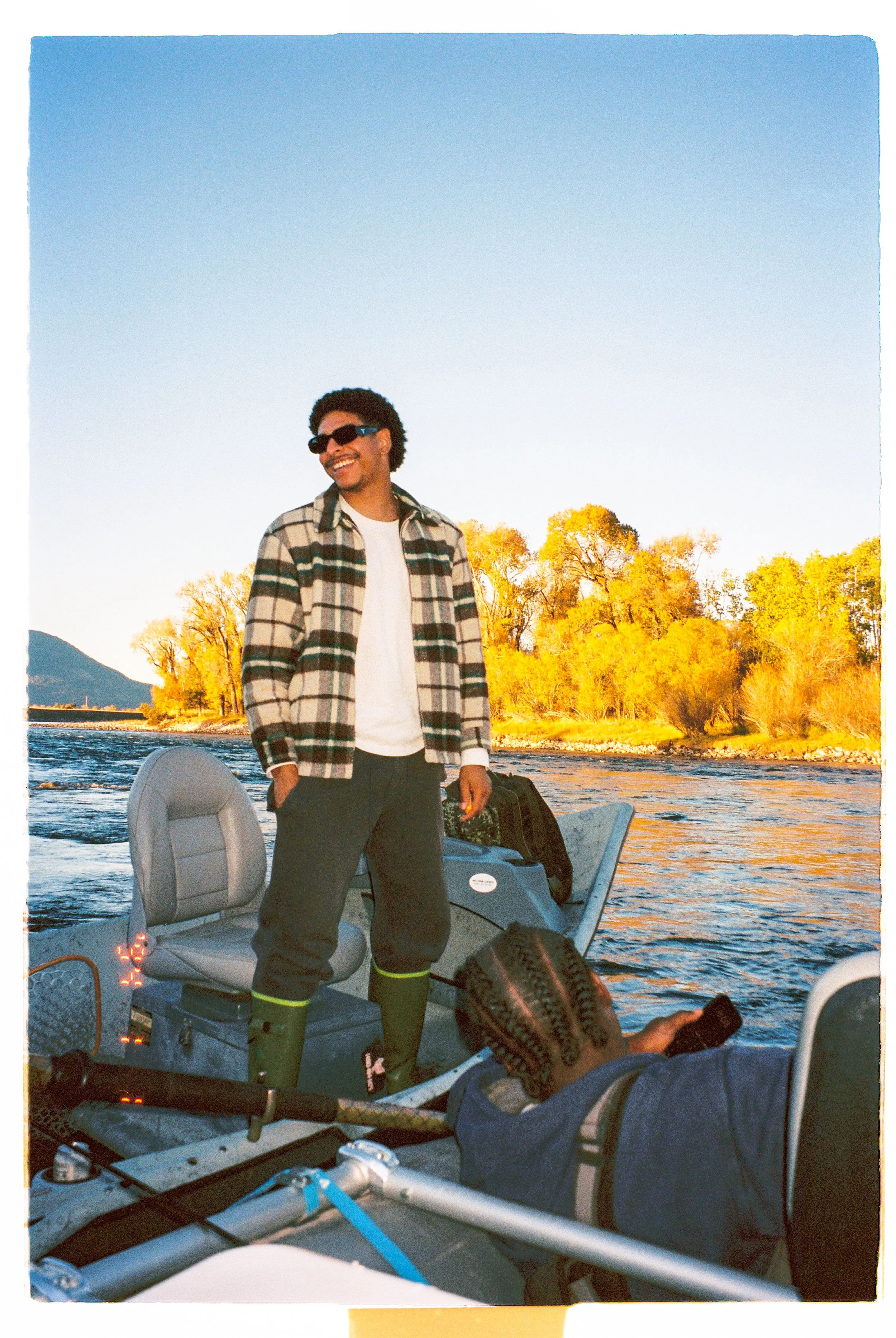 A man standing on a boat in a river, smiling with sunglasses, wearing a plaid shirt, white t-shirt, and dark pants, with a woman lying down on the boat and using a phone, surrounded by trees with autumn foliage and a mountain in the background.