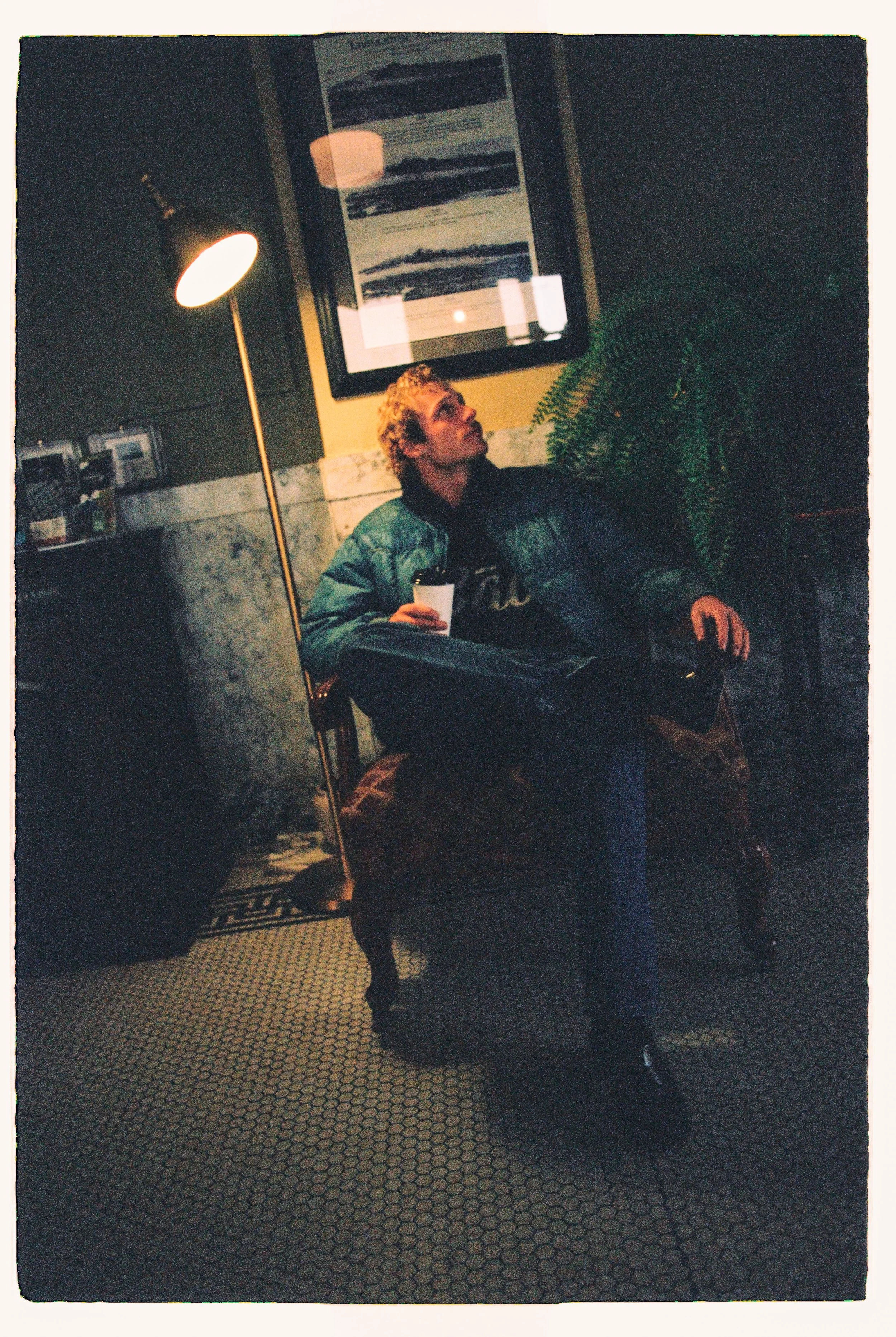 A person with short curly blonde hair sits in a dimly lit cafe, holding a cup, looking upward. They wear a green puffer jacket, black pants, and black shoes. Behind them is a picture frame on the wall, a green plant, and a standing lamp.