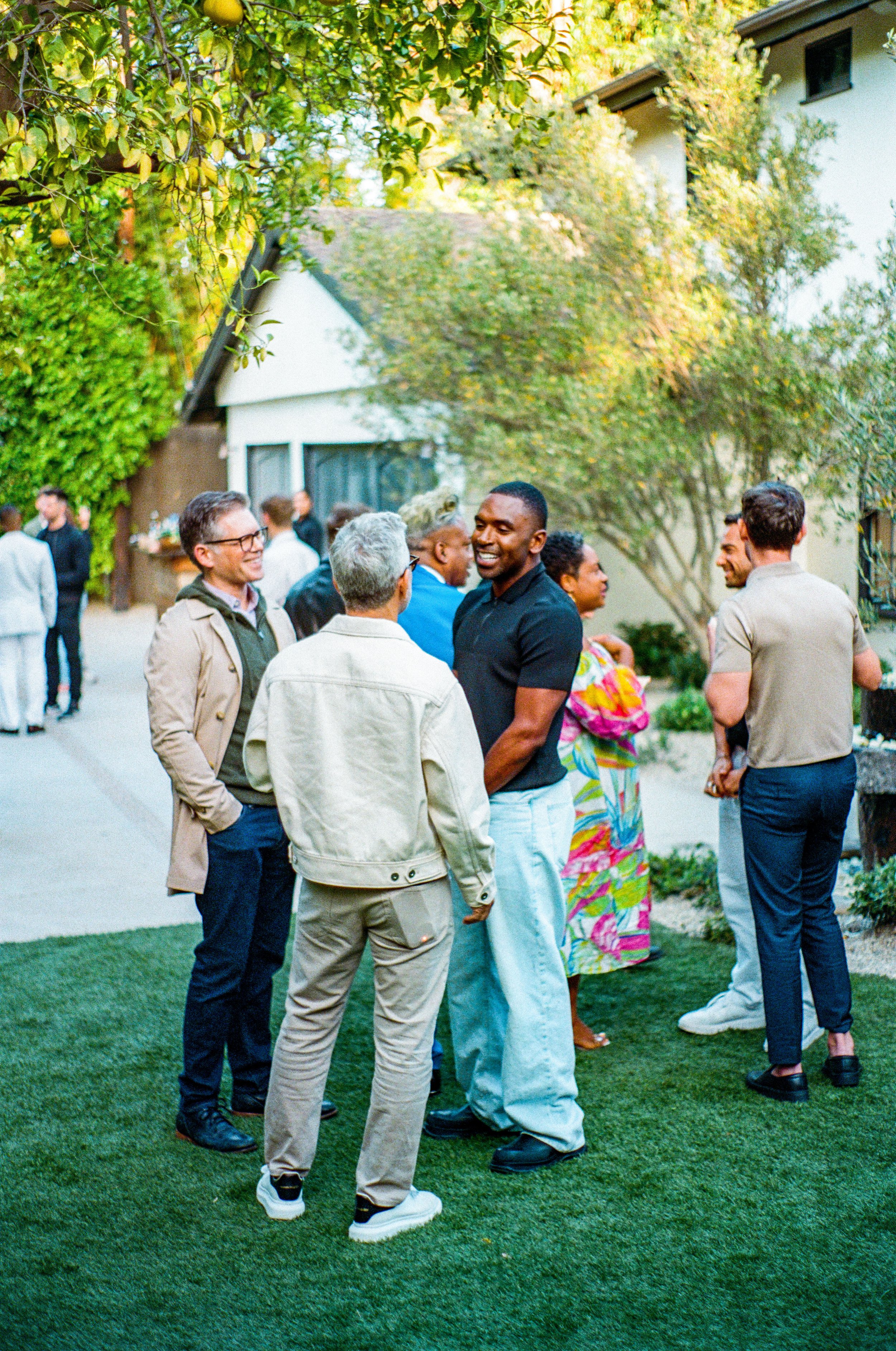 Group of people socializing outdoors in a garden or backyard, with trees and a house in the background, during daytime or early evening.