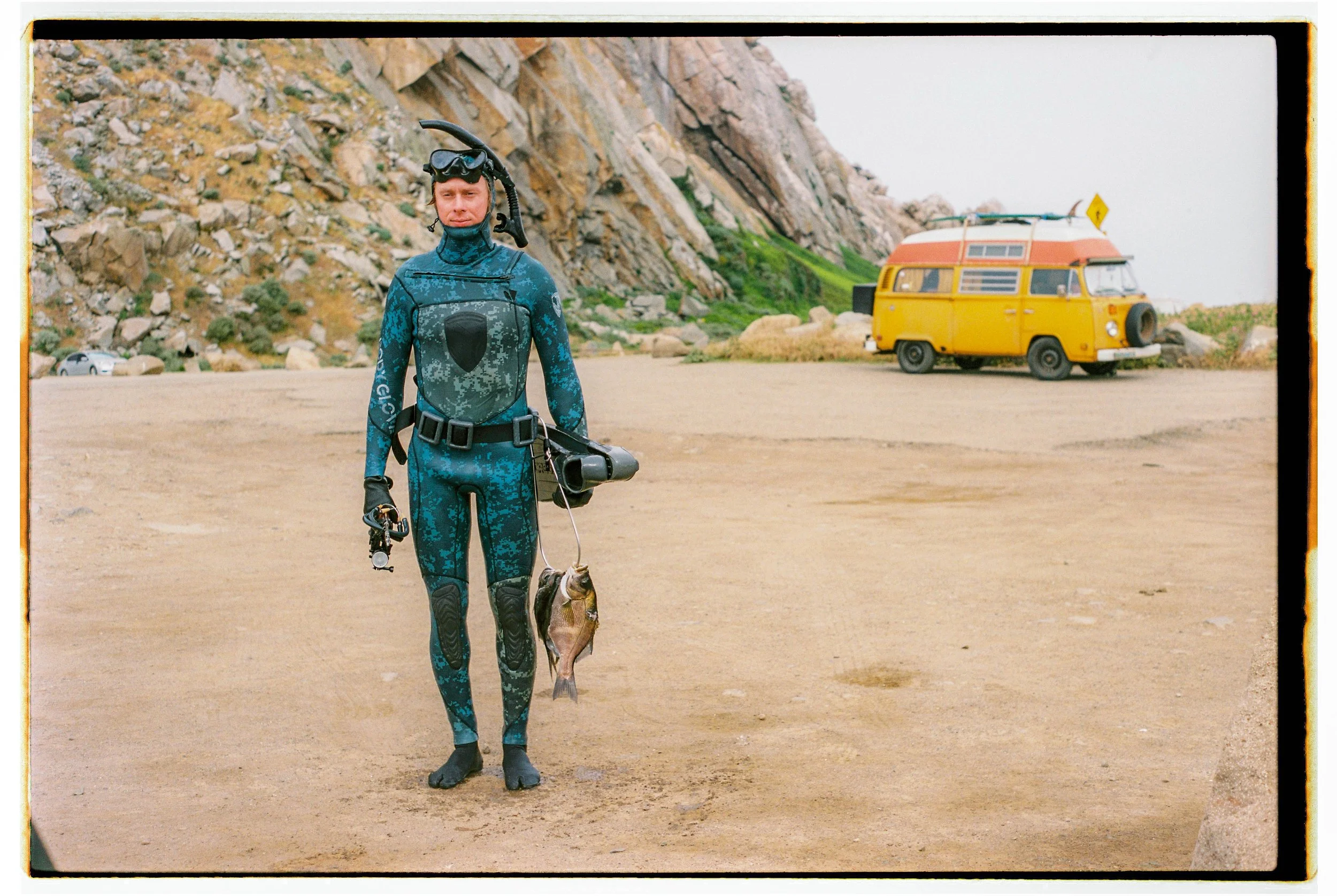 A person in wetsuit holding a fish with a yellow camper van in the background on a sandy beach with rocky cliffs.