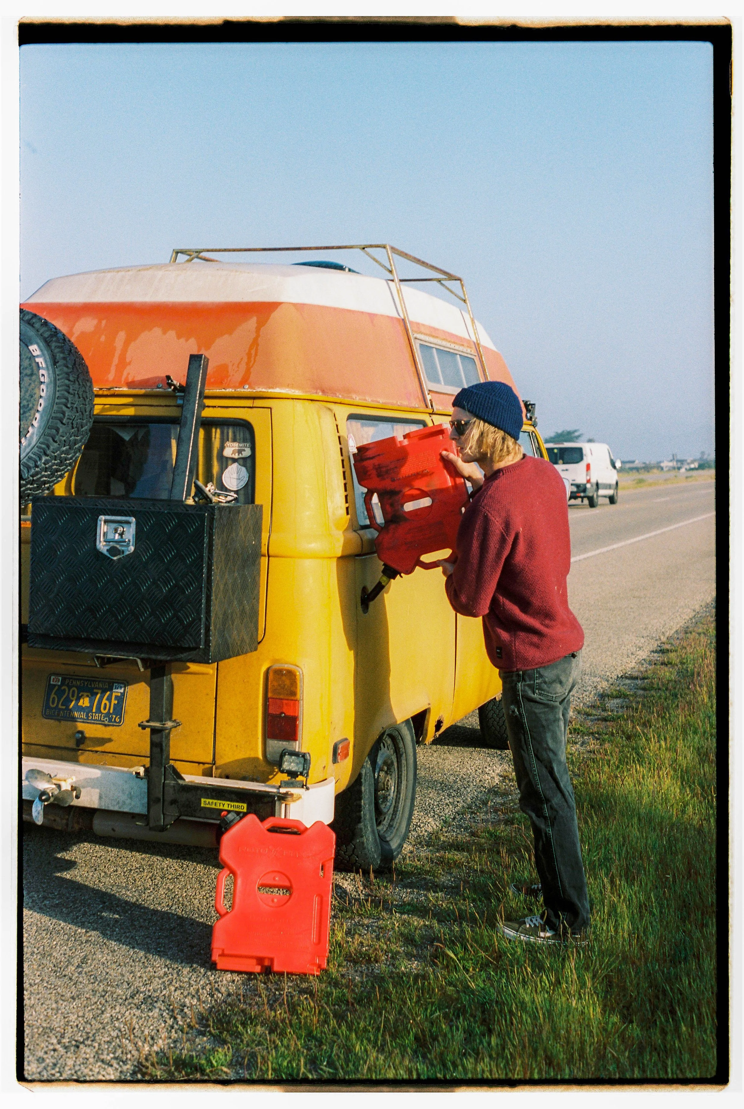 A person with blonde hair, wearing a blue beanie, sunglasses, a red sweater, and black pants, is fueling a yellow and orange vintage camper van with a red gas canister near a highway. There's a red gas canister on the ground and a black toolbox mount