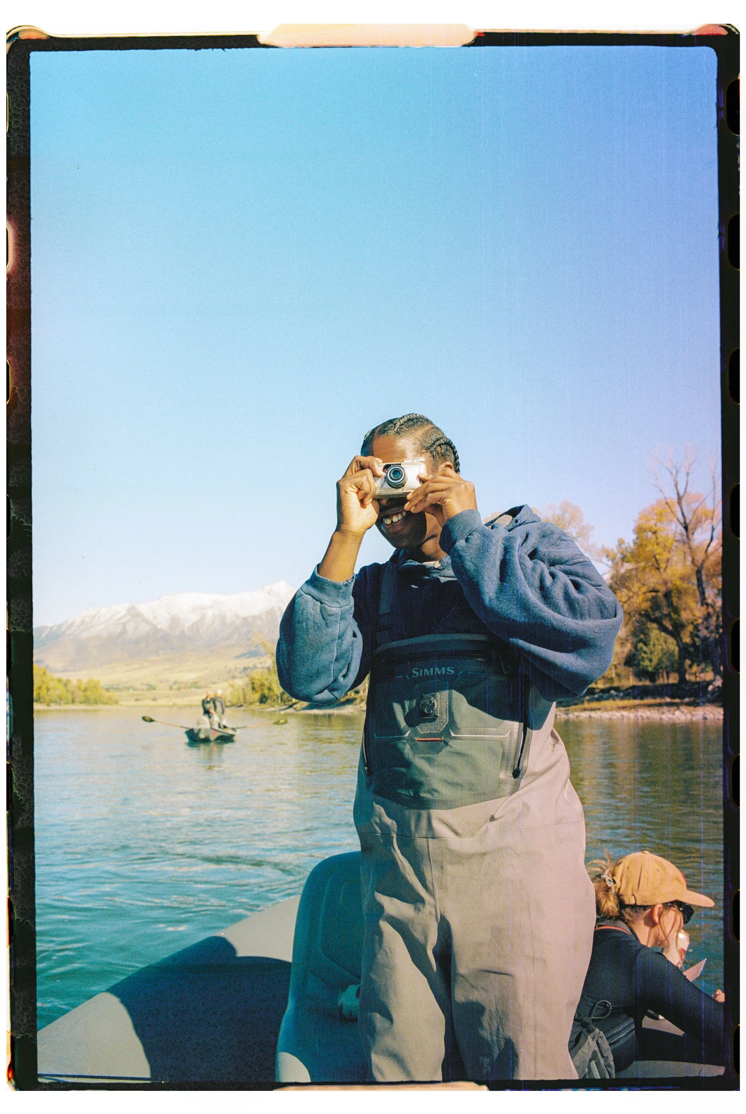 Person taking a photograph on a boat, smiling, with mountains and a river in the background, and another person sitting on the boat.