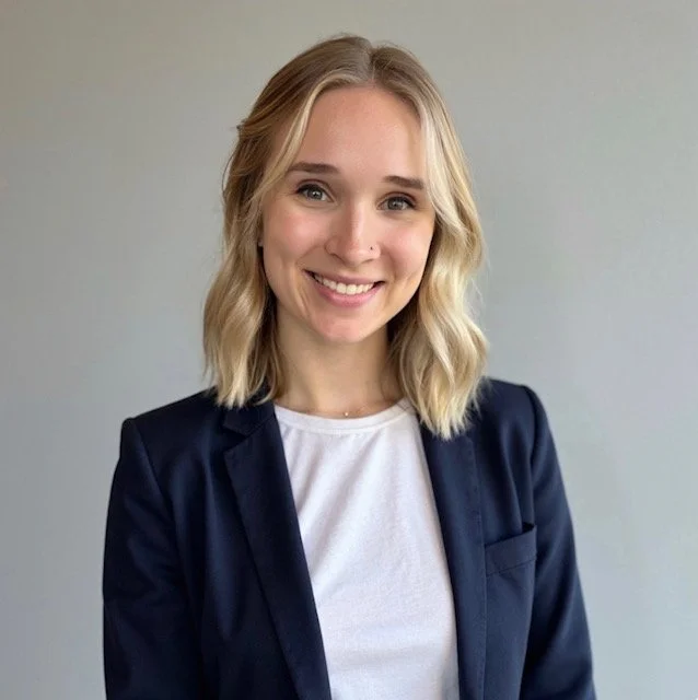 A young woman with blonde hair in loose waves, smiling, wearing a navy blazer and white t-shirt, standing against a plain gray background.