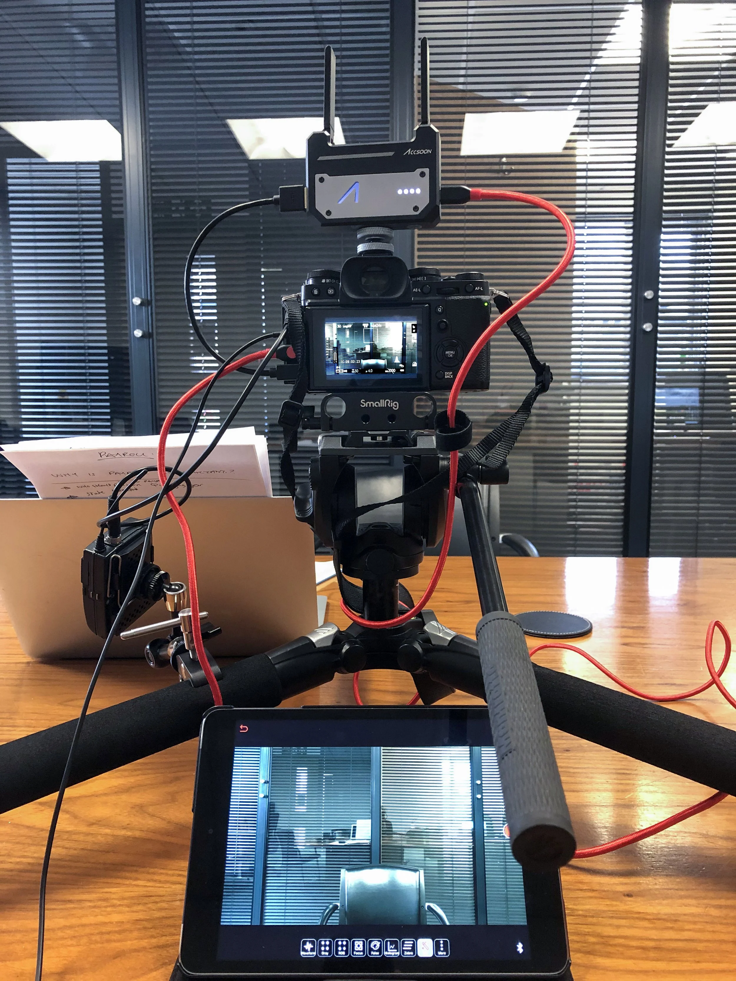 A camera on a tripod set up on a wooden table recording a room with glass walls and closed blinds.