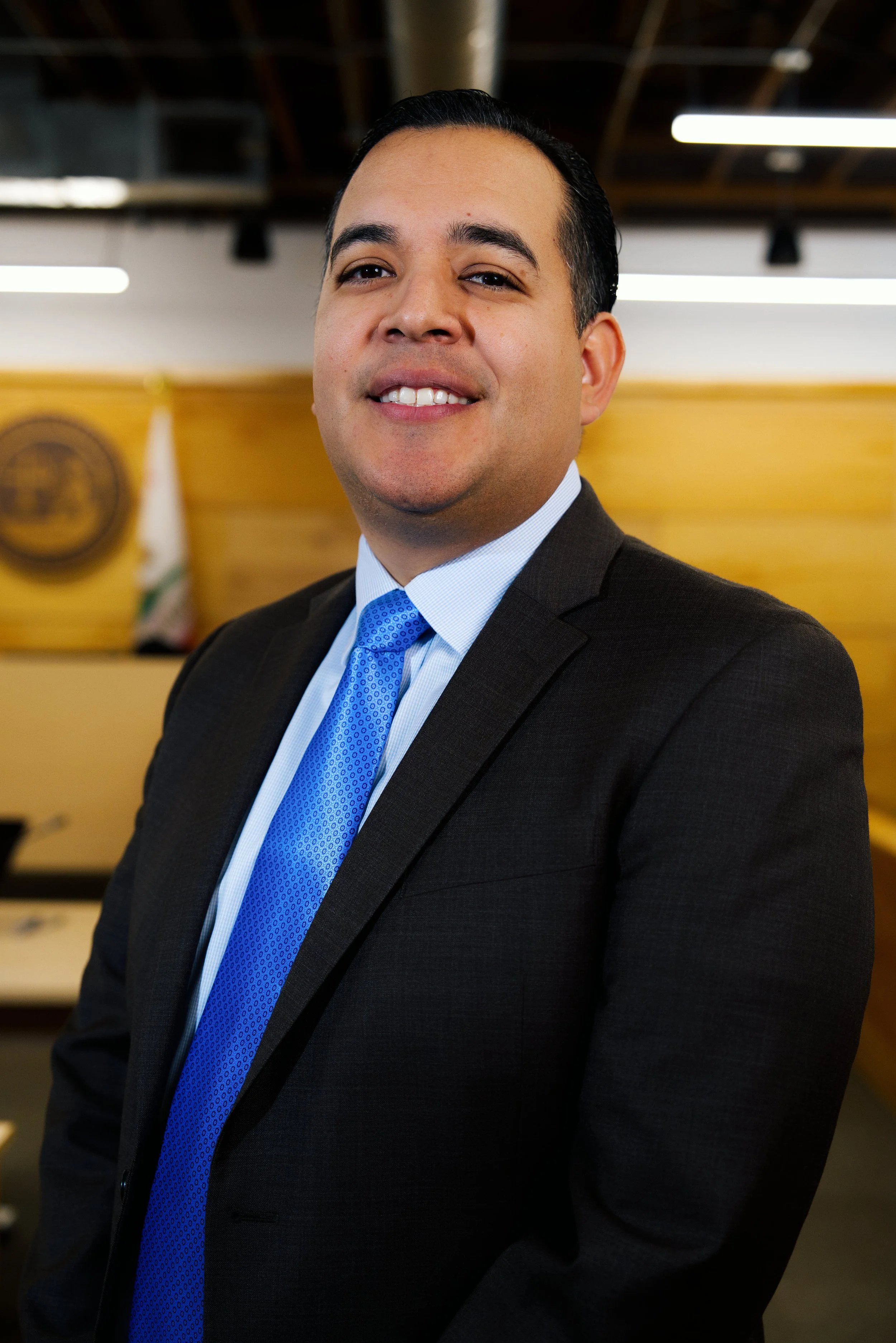 Hombre de negocios con traje oscuro y corbata azul, sonriendo en una oficina formal.