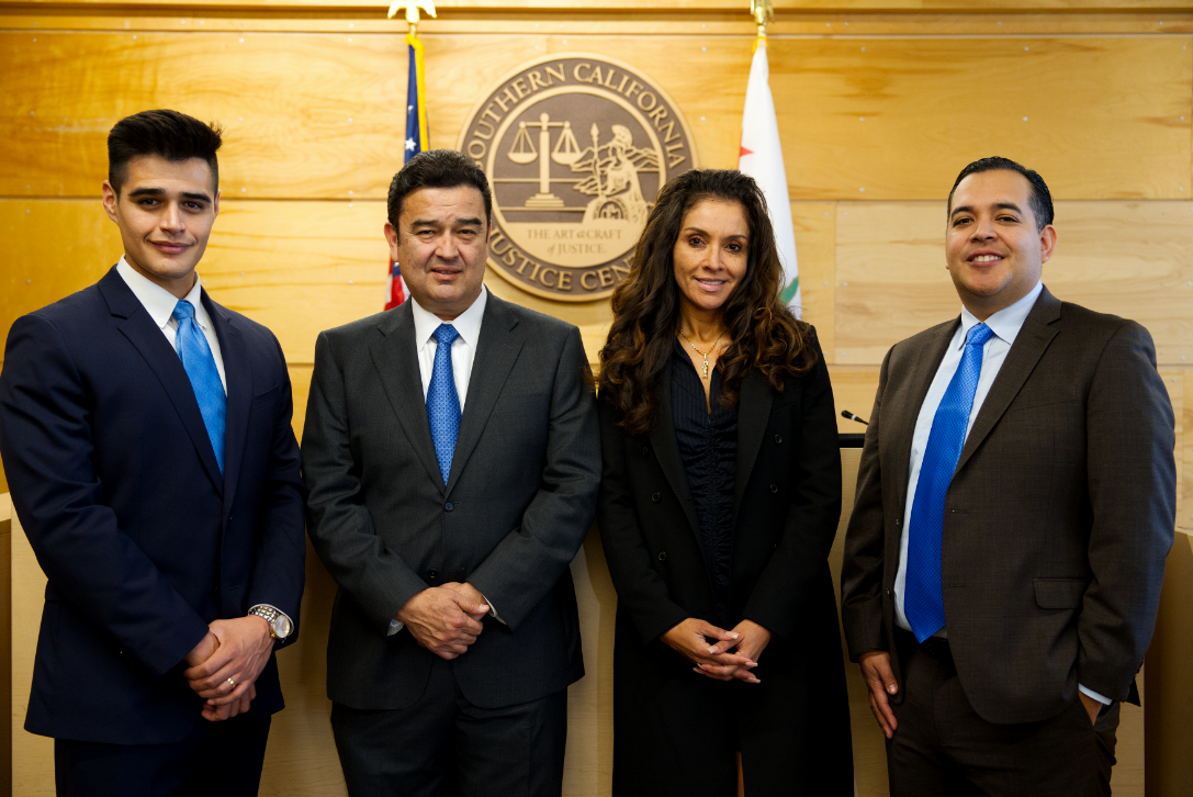 Cuatro personas vestidas con traje formal posando en una sala con el escudo de la Corte de Justicia del Sur de California en la pared y banderas de EE.UU. y California en el fondo.