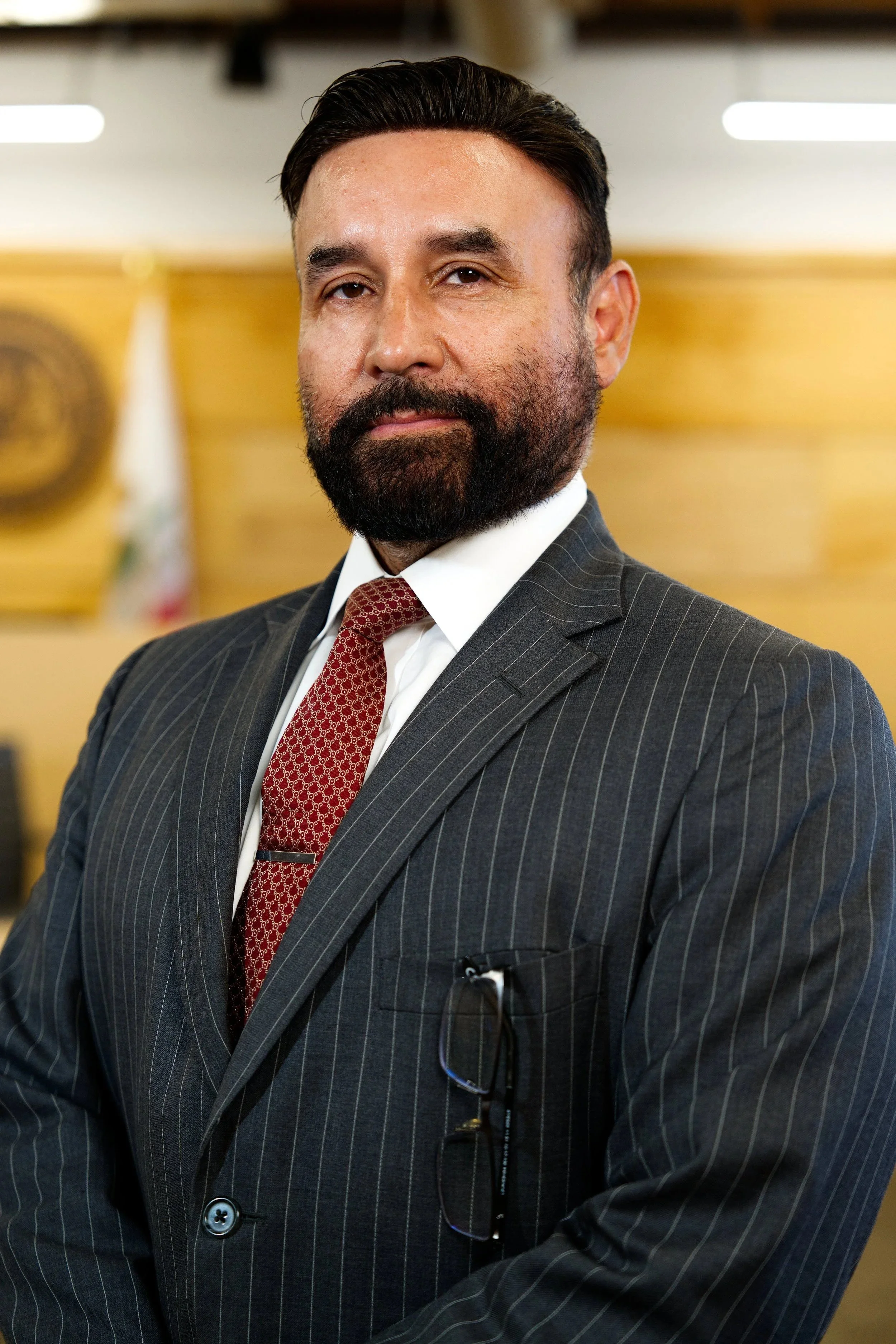 Hombre con barba y sonrisa, usando traje oscuro con raya y corbata roja, en una oficina o sala formal.