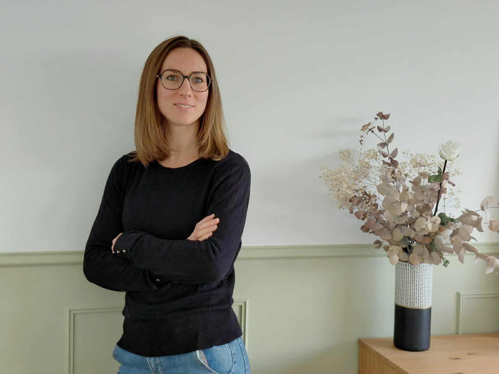 Une femme aux cheveux bruns, portant des lunettes noires et une chemise noire, se tient avec les bras croisés à côté d'un vase de fleurs séchées sur un meuble en bois, dans une pièce avec un mur blanc et un panneau de couleur vert clair.