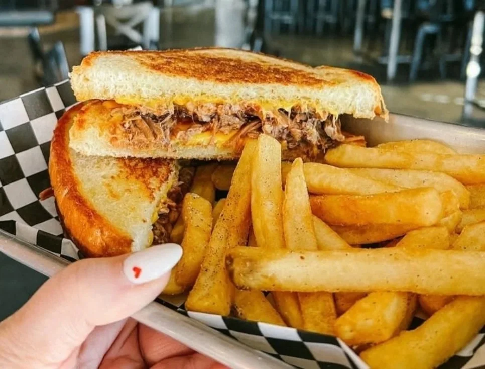 Half of a toasted cheeseburger with shredded beef and melted cheese, served with French fries on a checkered paper-lined tray.