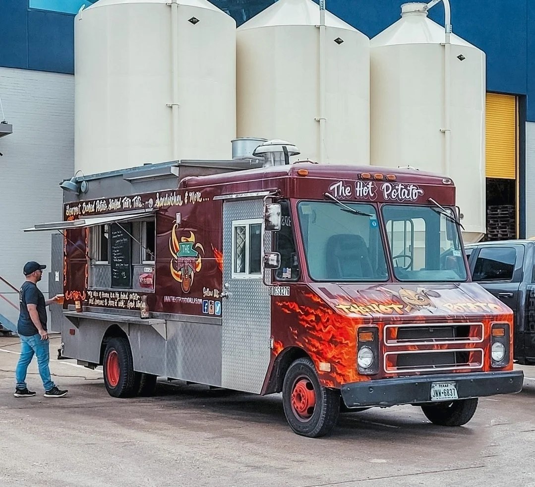 Food truck named 'The Hot Potato' parked outside with a man standing nearby, in front of large white storage tanks.
